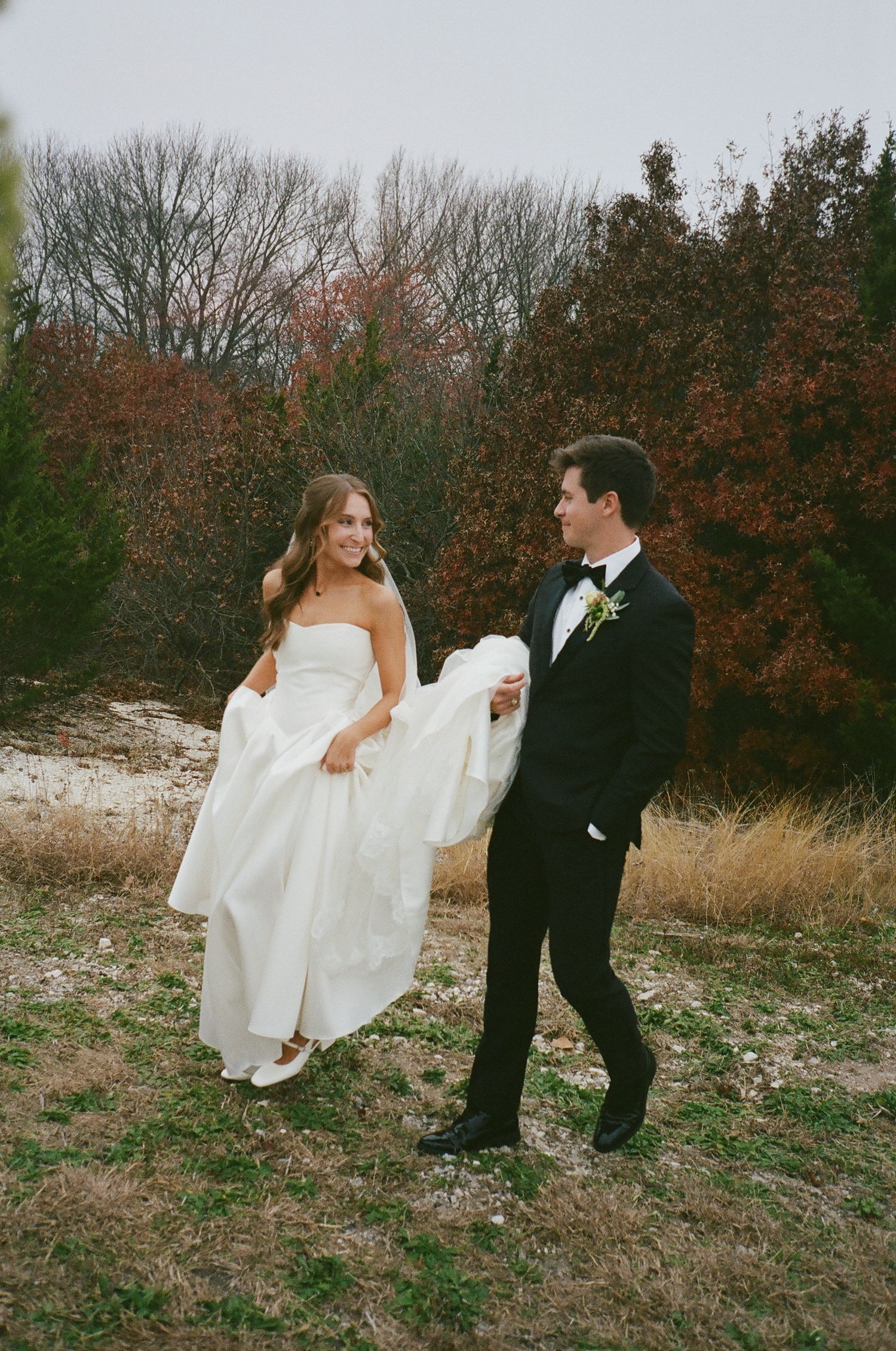 Smiling bride in strapless gown and groom in black tuxedo walking outdoors in autumn.