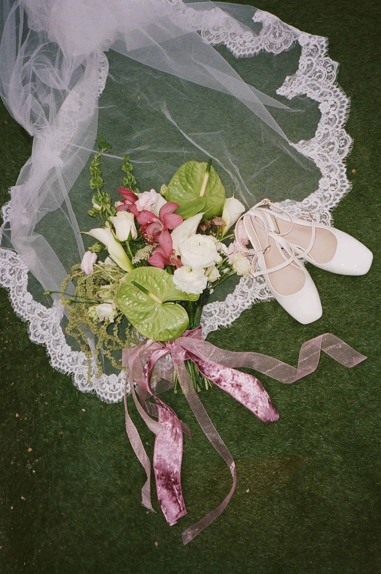Bridal flat shoes, lace veil, and pink-tied bouquet with anthurium flowers on grass.