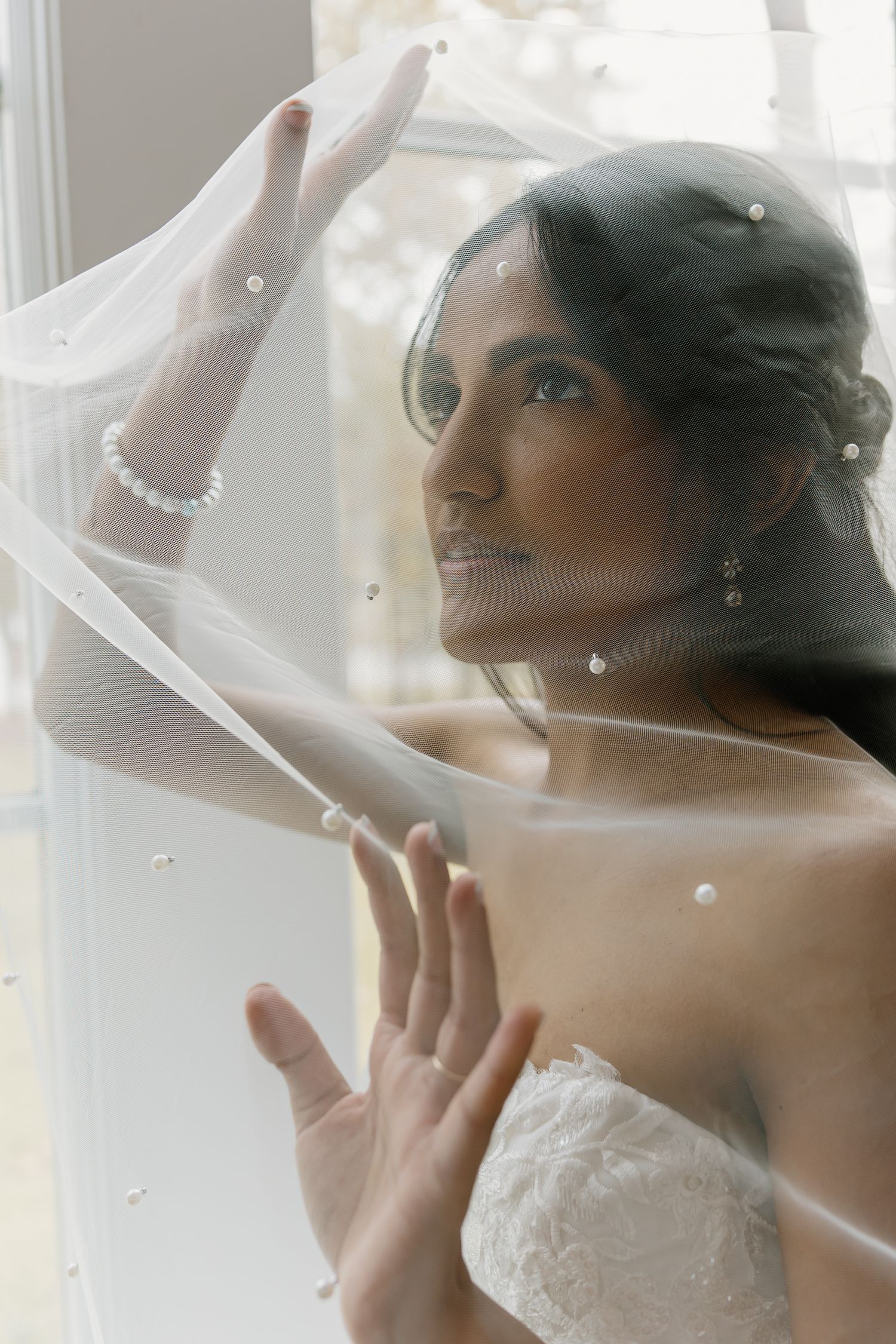 Stunning bride in lace strapless gown holds pearl-adorned veil while gazing through a bright window.
