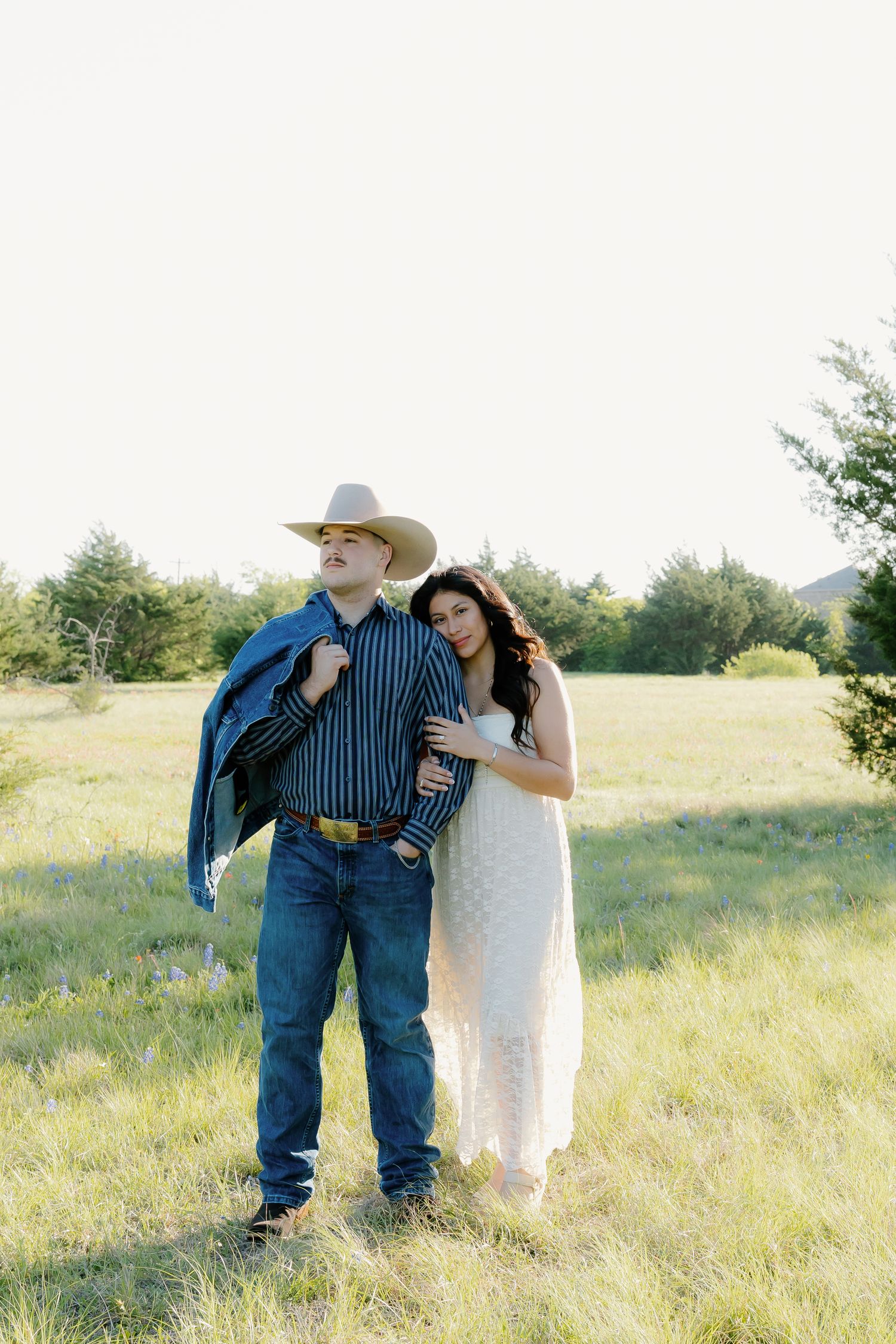 Couple posing in sunlit field, man in cowboy hat and jeans, woman in white lace dress.