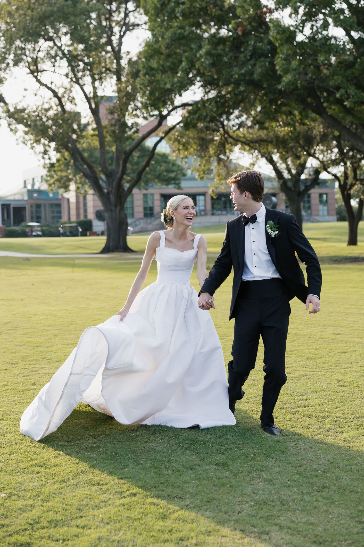 Joyful bride and groom running on green lawn, bride laughing in white ball gown, groom in black tuxedo.