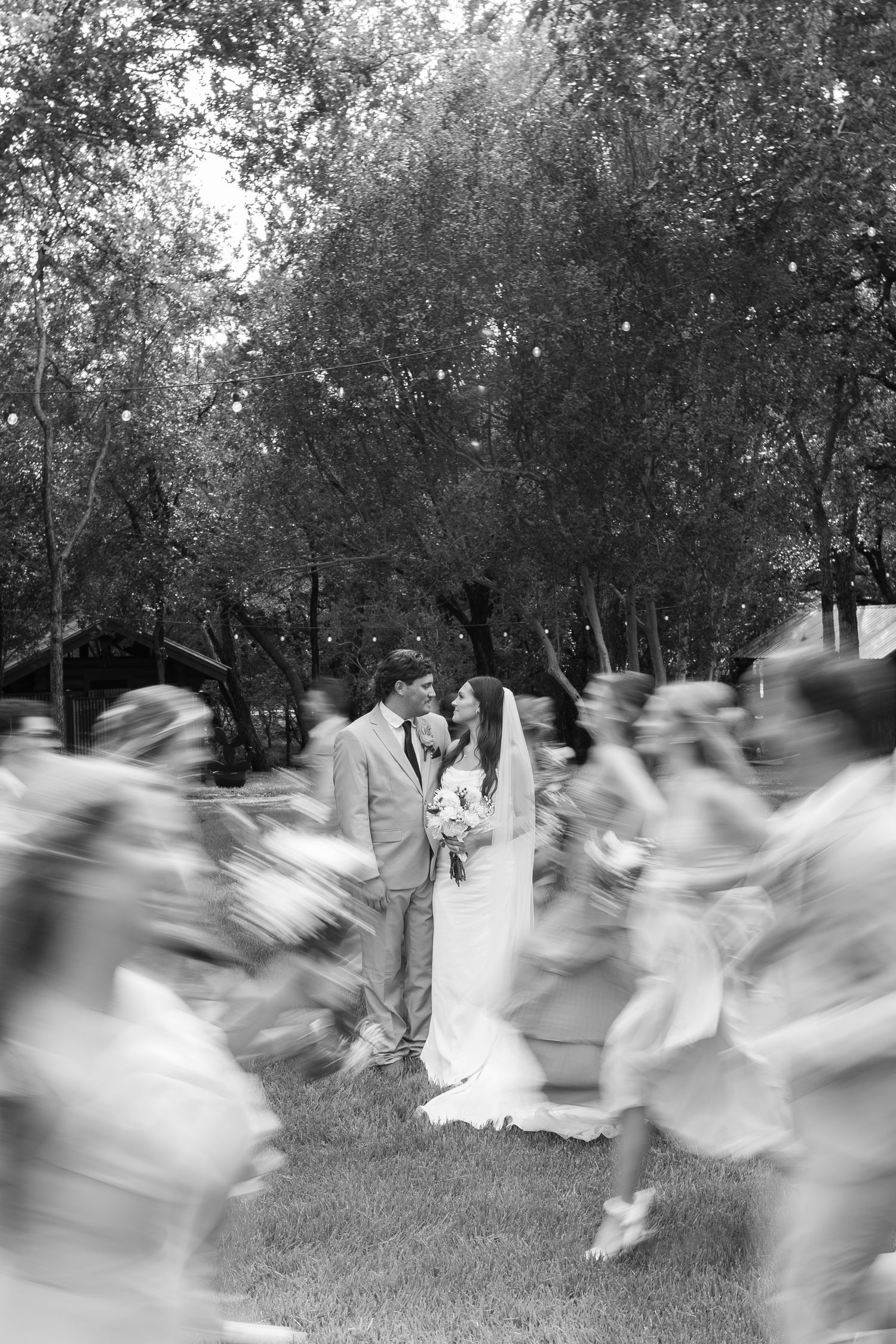Black and white photo of bride and groom standing still as wedding party moves around them.