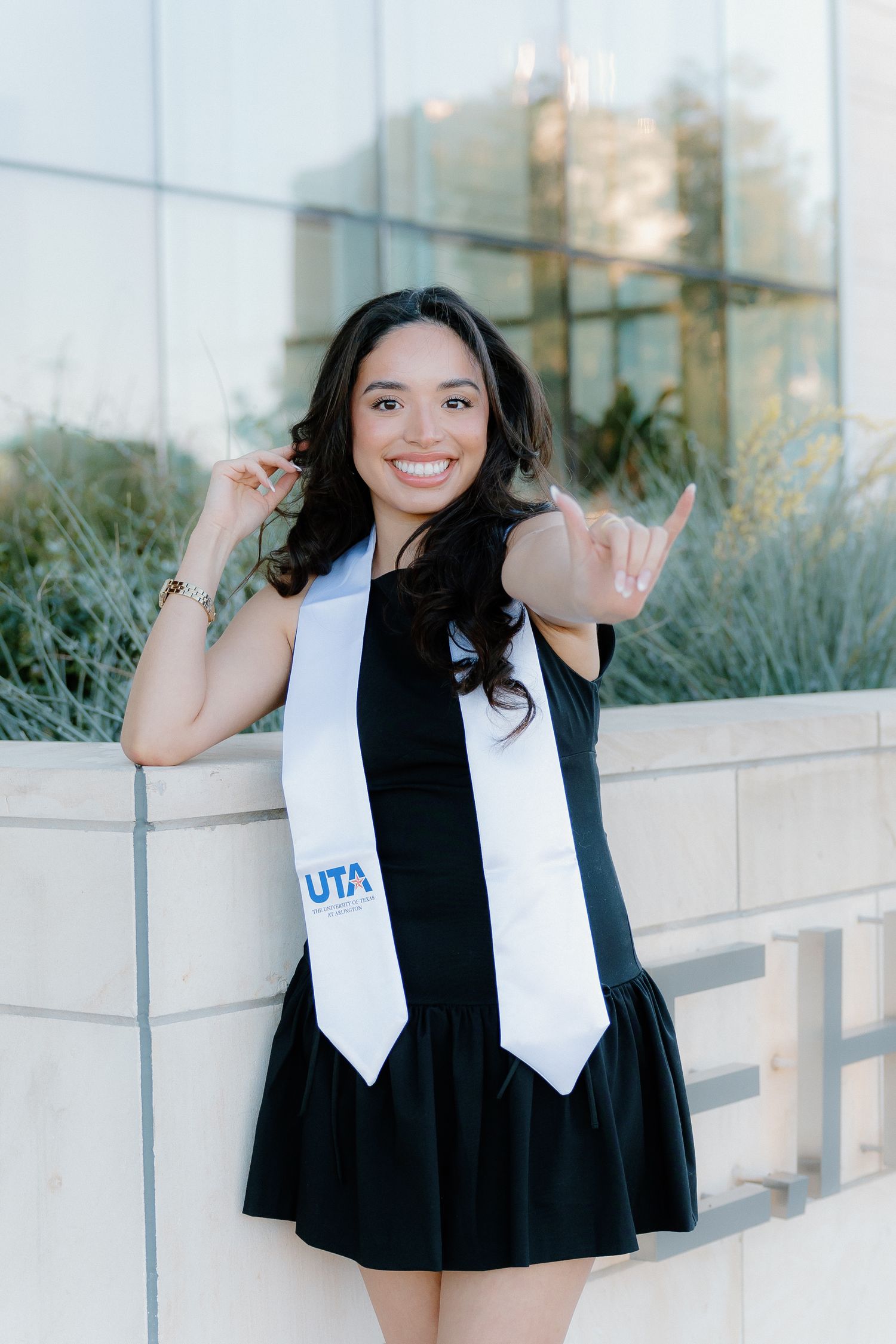 Smiling UTA graduate in black dress with white graduation stole making school spirit hand gestures outdoors.