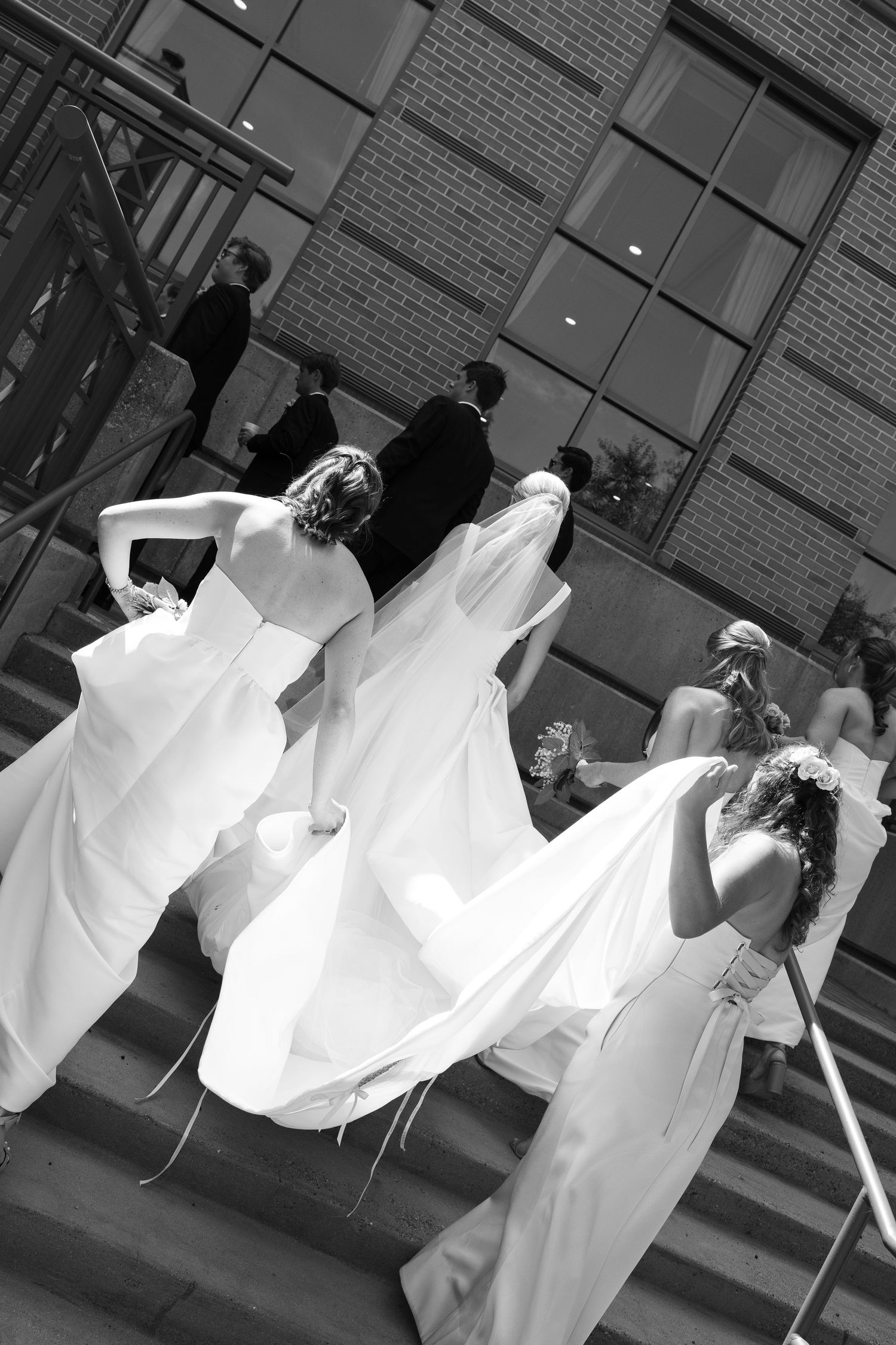 Black and white photo of bridesmaids in white gowns helping bride up stairs at wedding venue.