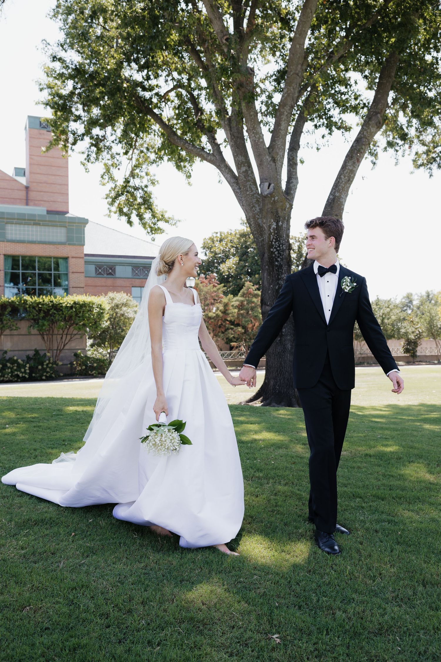 Bride and groom holding hands walking on green lawn with large tree in background.