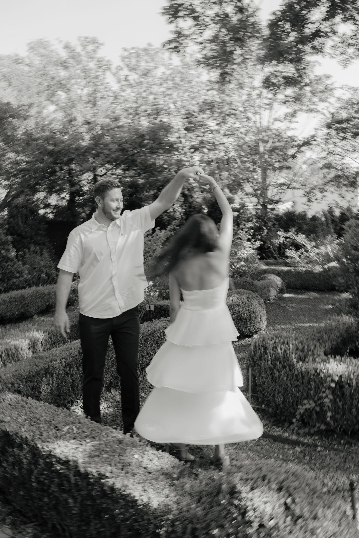Black and white photo of a couple dancing in a garden, groom spinning bride in tiered white dress.