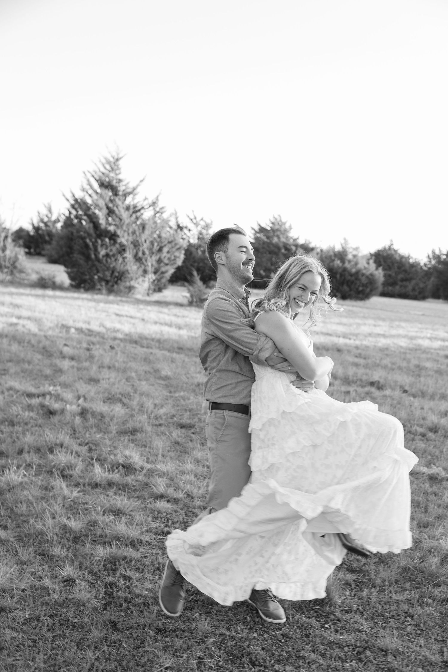 Groom lifting bride in wedding dress outdoors in black and white photo, both laughing joyfully in a field.