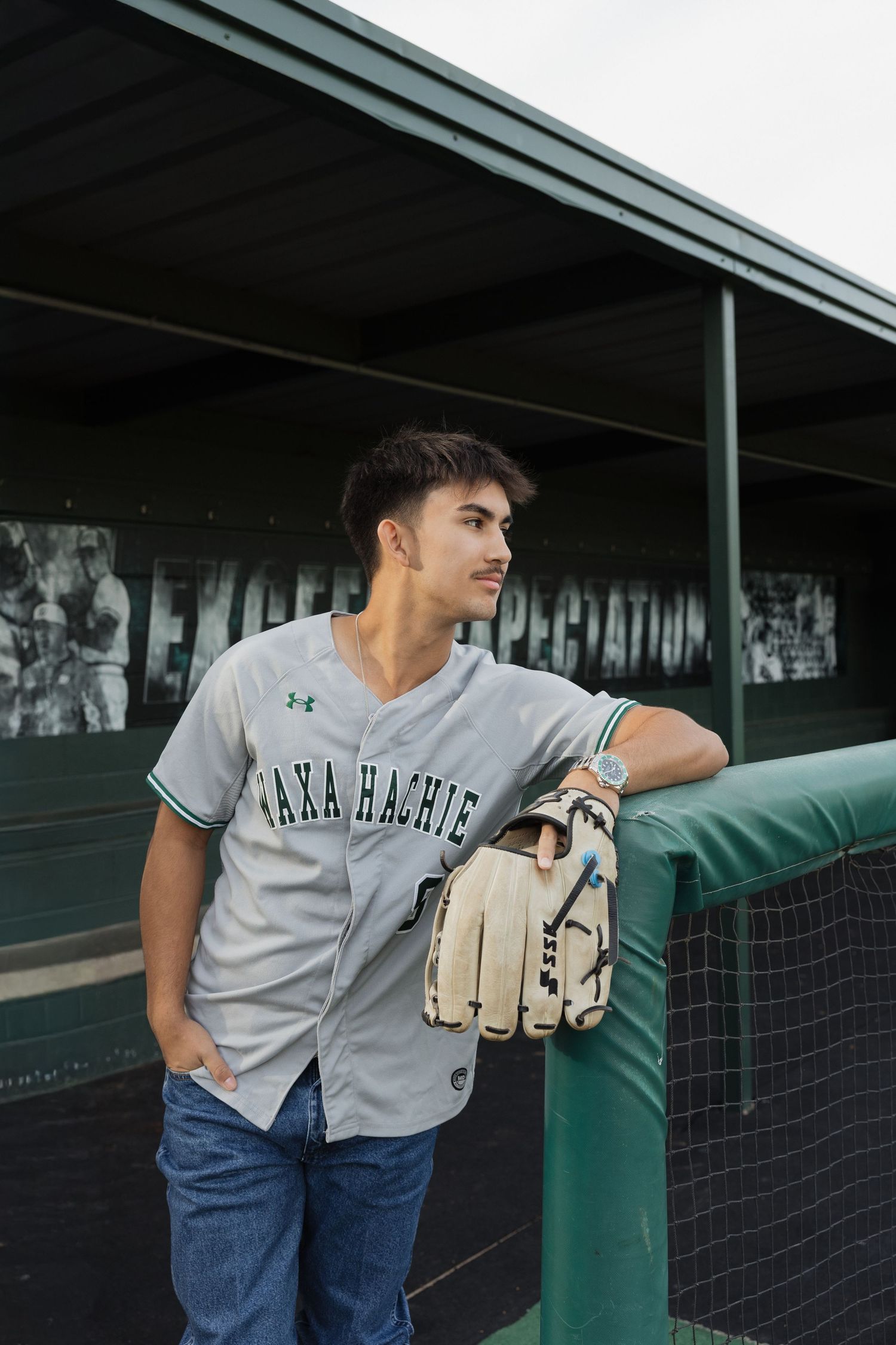 Young baseball player in Waxahachie jersey holding glove, leaning on dugout fence at baseball field.
