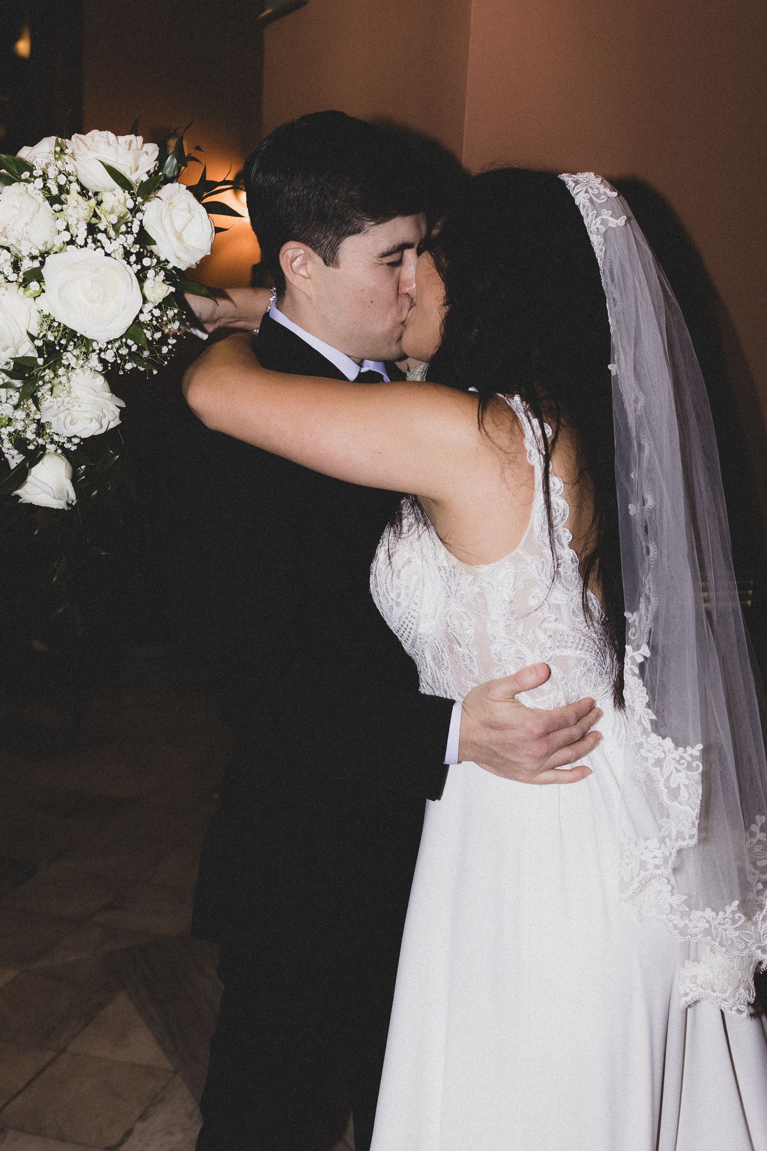 Newlywed couple sharing first kiss surrounded by white roses at evening wedding ceremony.
