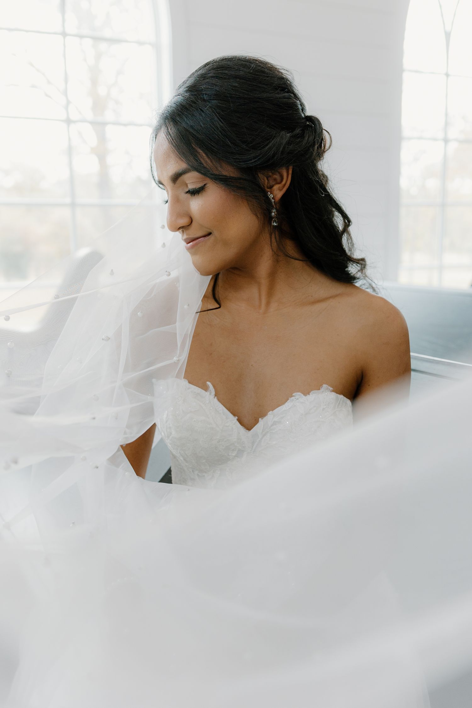 Radiant bride in strapless lace gown with pearl veil smiles softly in bright natural window light.