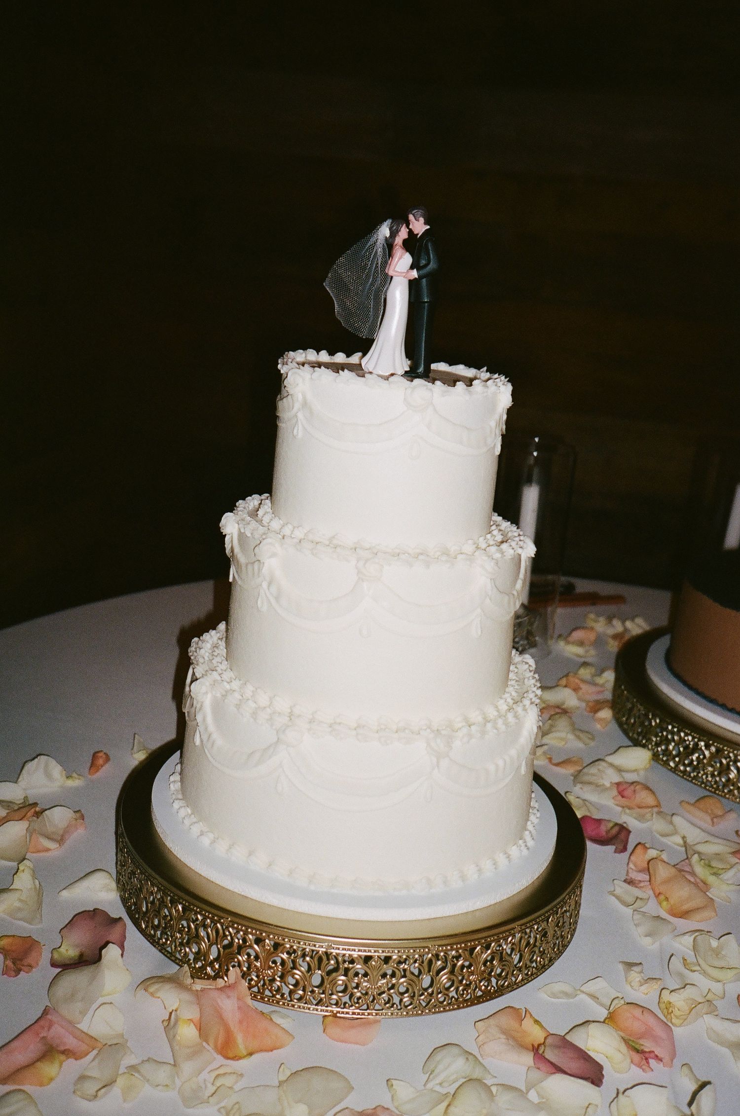 Three-tier white wedding cake with piped details and bride and groom topper on gold stand.