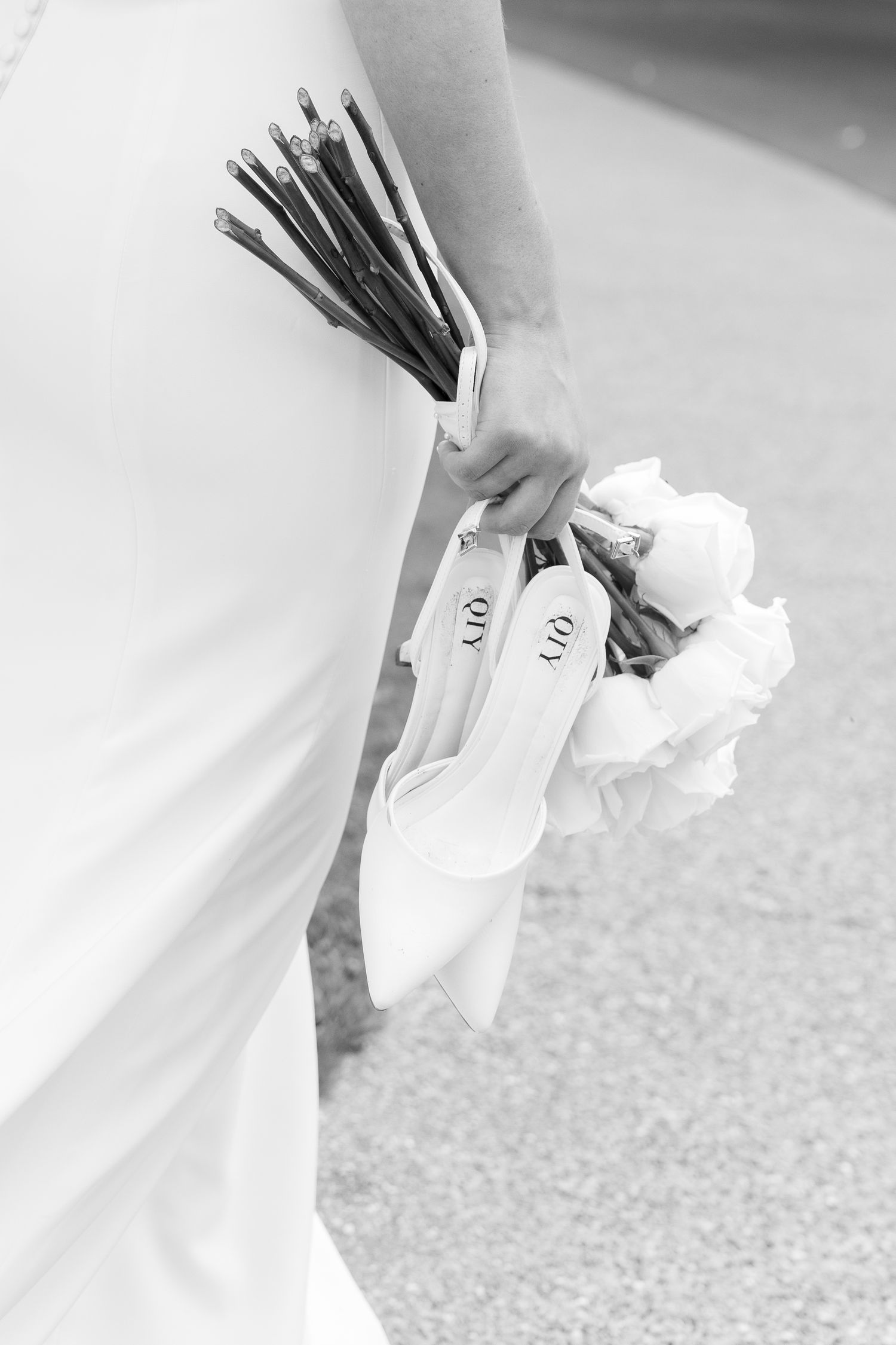 Black and white image of bride holding white heels and bouquet with dark ribbon stems.