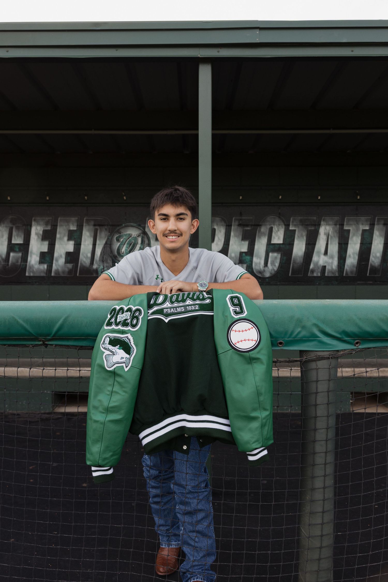 Smiling teen holds green letterman jacket with baseball patch outside dugout wearing Braves shirt and jeans.