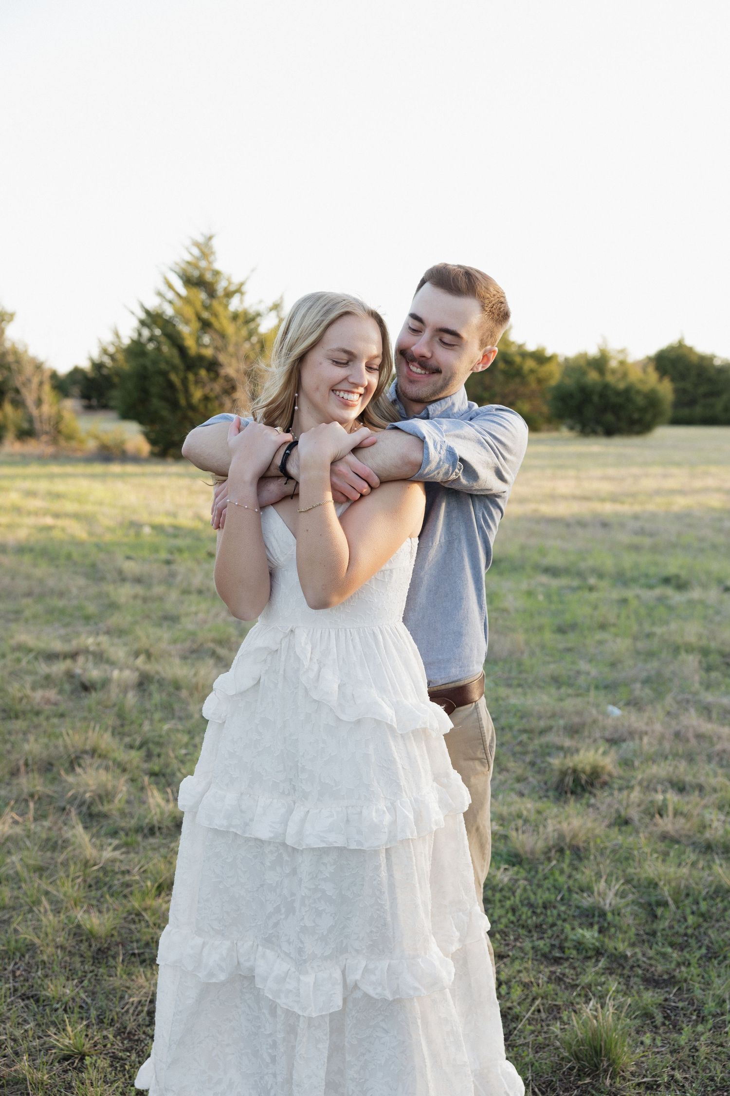 A groom embraces his bride in a white ruffled dress in a sunlit field, both smiling joyfully.