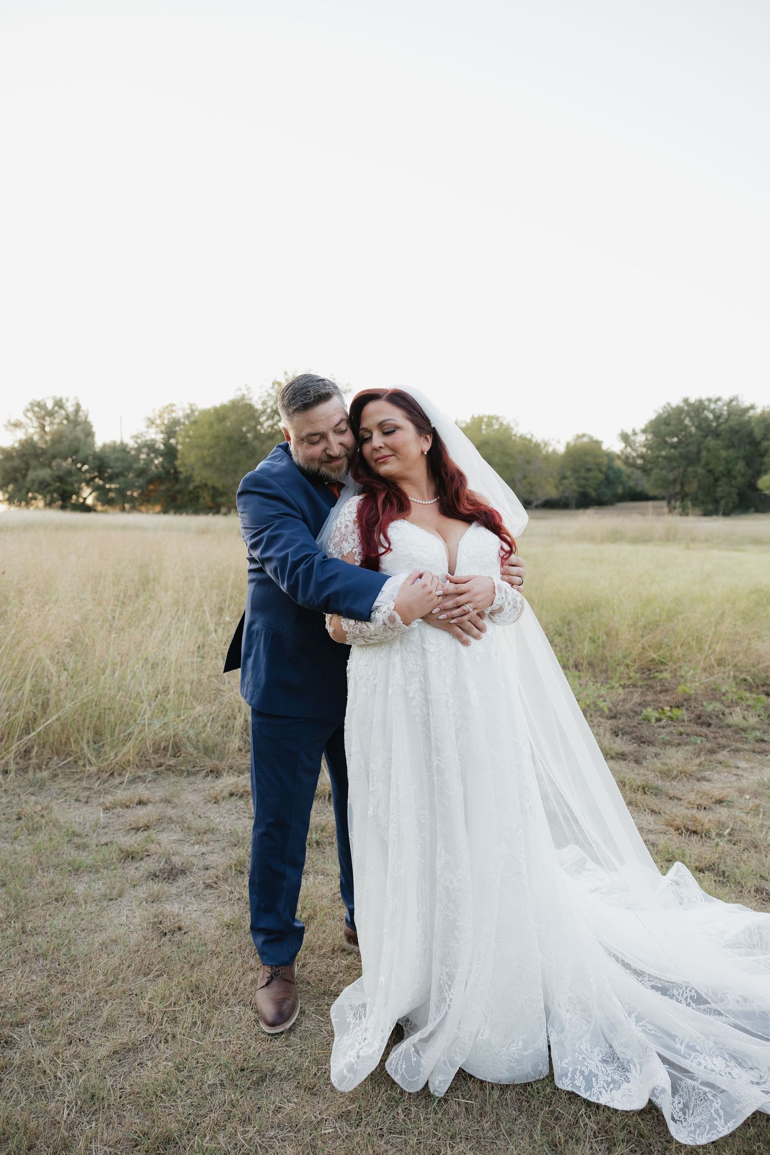 Groom embraces bride in lace wedding gown from behind in a sunlit open field.