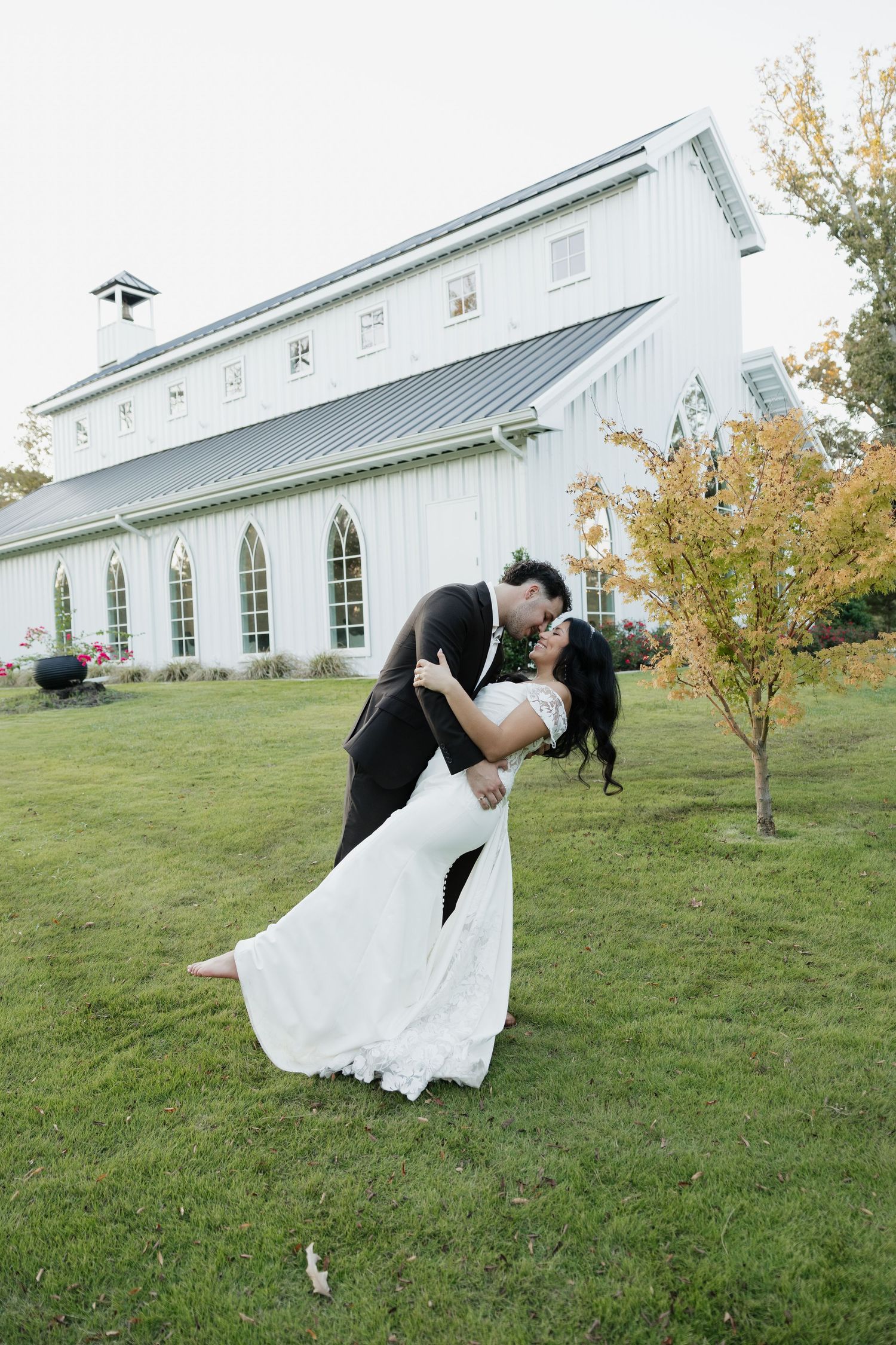 Groom dips bride in a romantic kiss outside a white barn wedding venue surrounded by autumn trees.