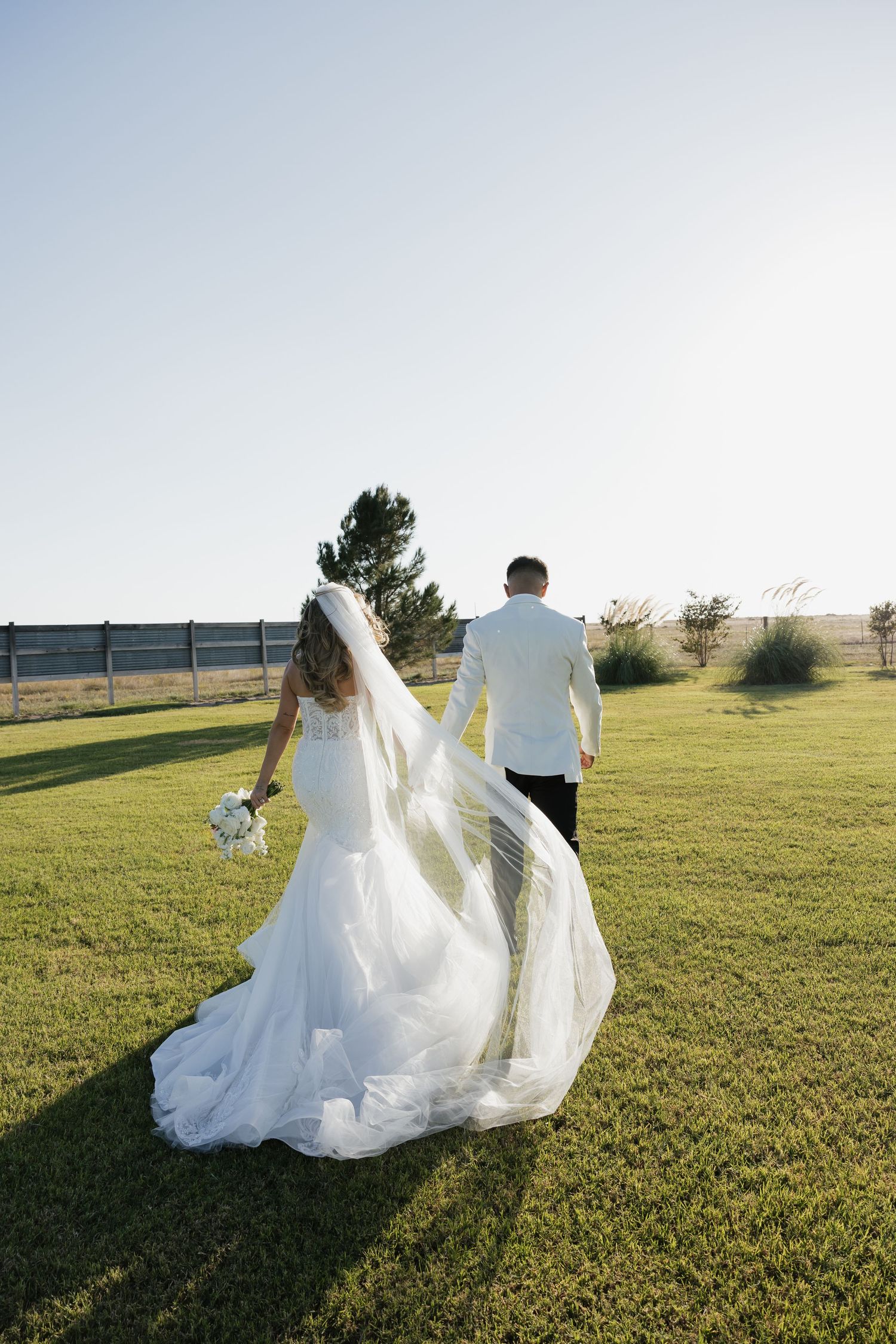 Bride and groom walk hand in hand across green lawn, wedding veil flowing in the breeze.