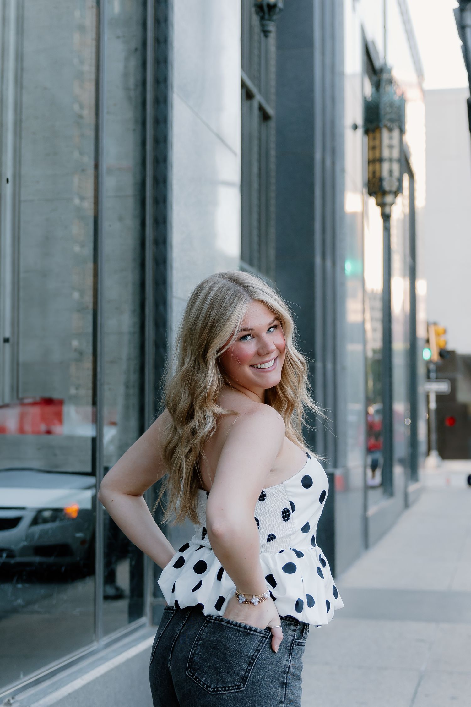 Smiling blonde woman in white polka dot strapless top and jeans posing on an urban city sidewalk.