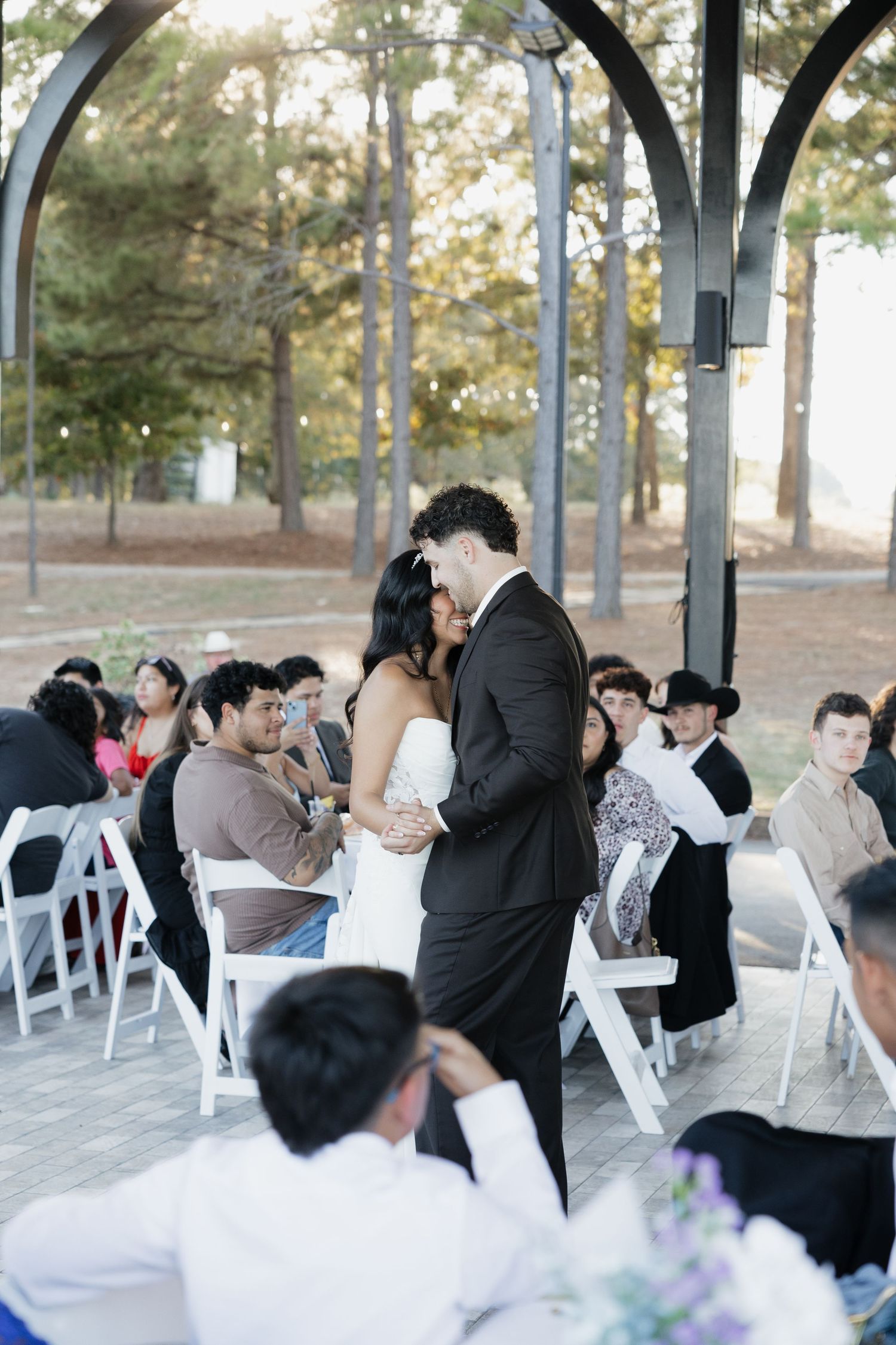 Bride and groom sharing first kiss at outdoor gazebo wedding ceremony with seated guests.