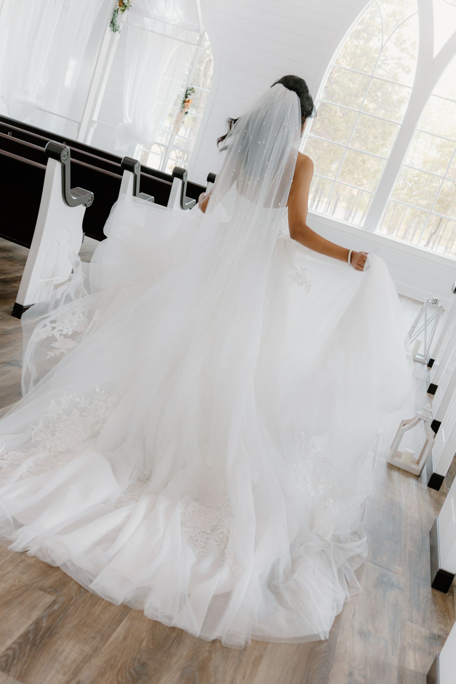 Bride in full ballgown with lace train and veil walks down chapel aisle toward arched window.
