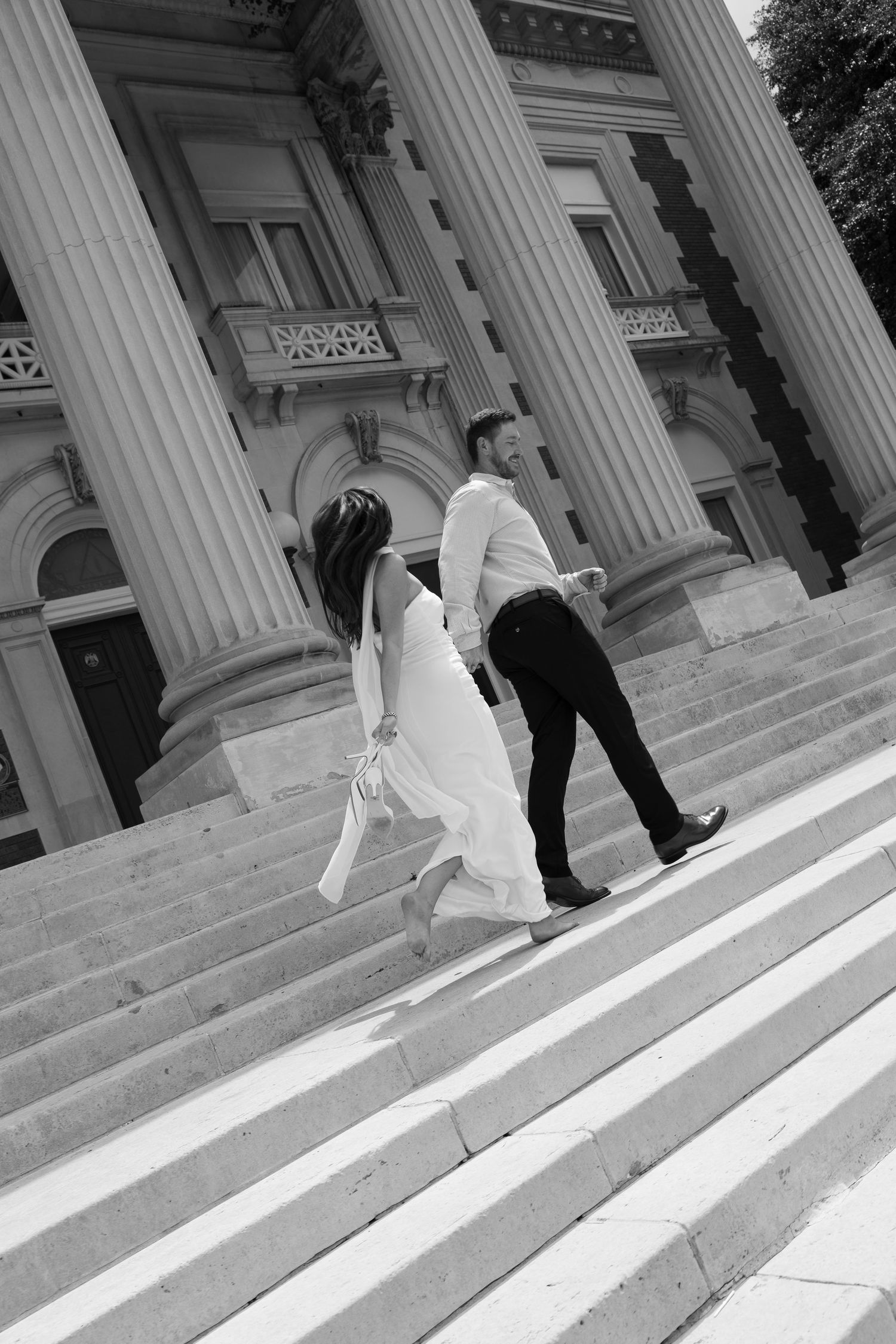Black and white photo of a couple holding hands walking up grand stone steps of a classical building with tall columns.