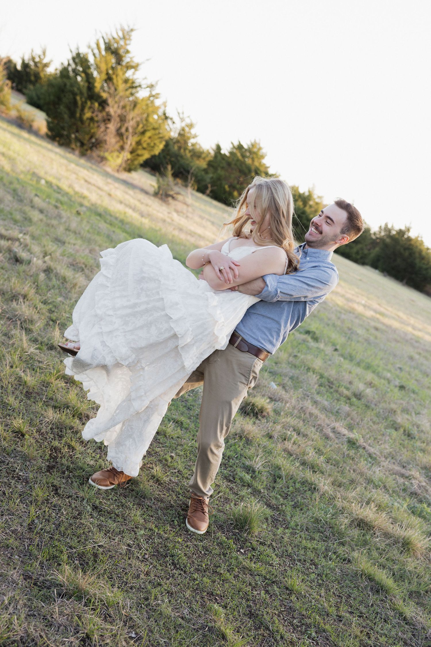 Groom lifting bride in white wedding dress outdoors on grassy hillside, both smiling joyfully at golden hour.
