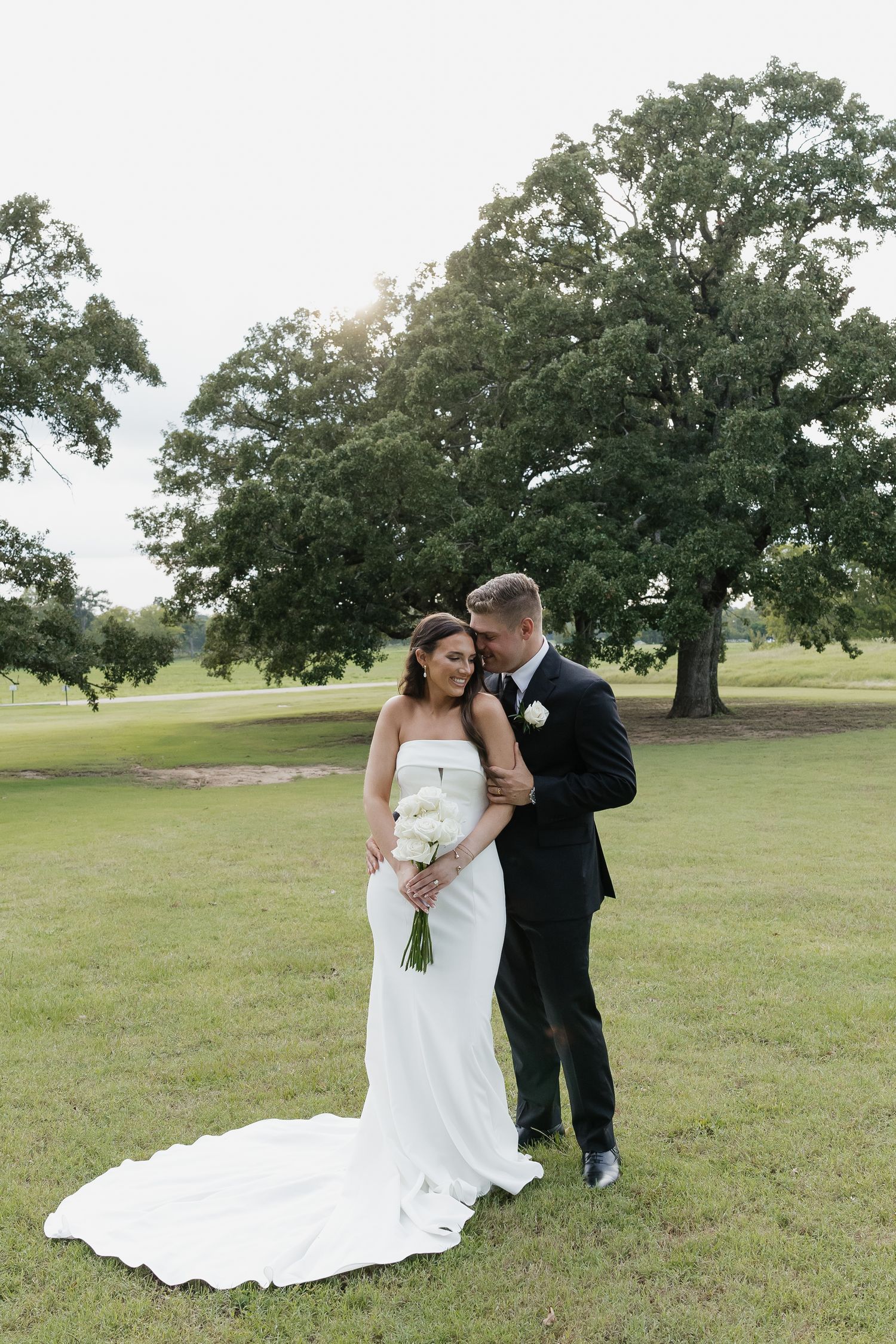 Groom tenderly embraces bride in fitted white gown while standing on lush green lawn near oak tree.