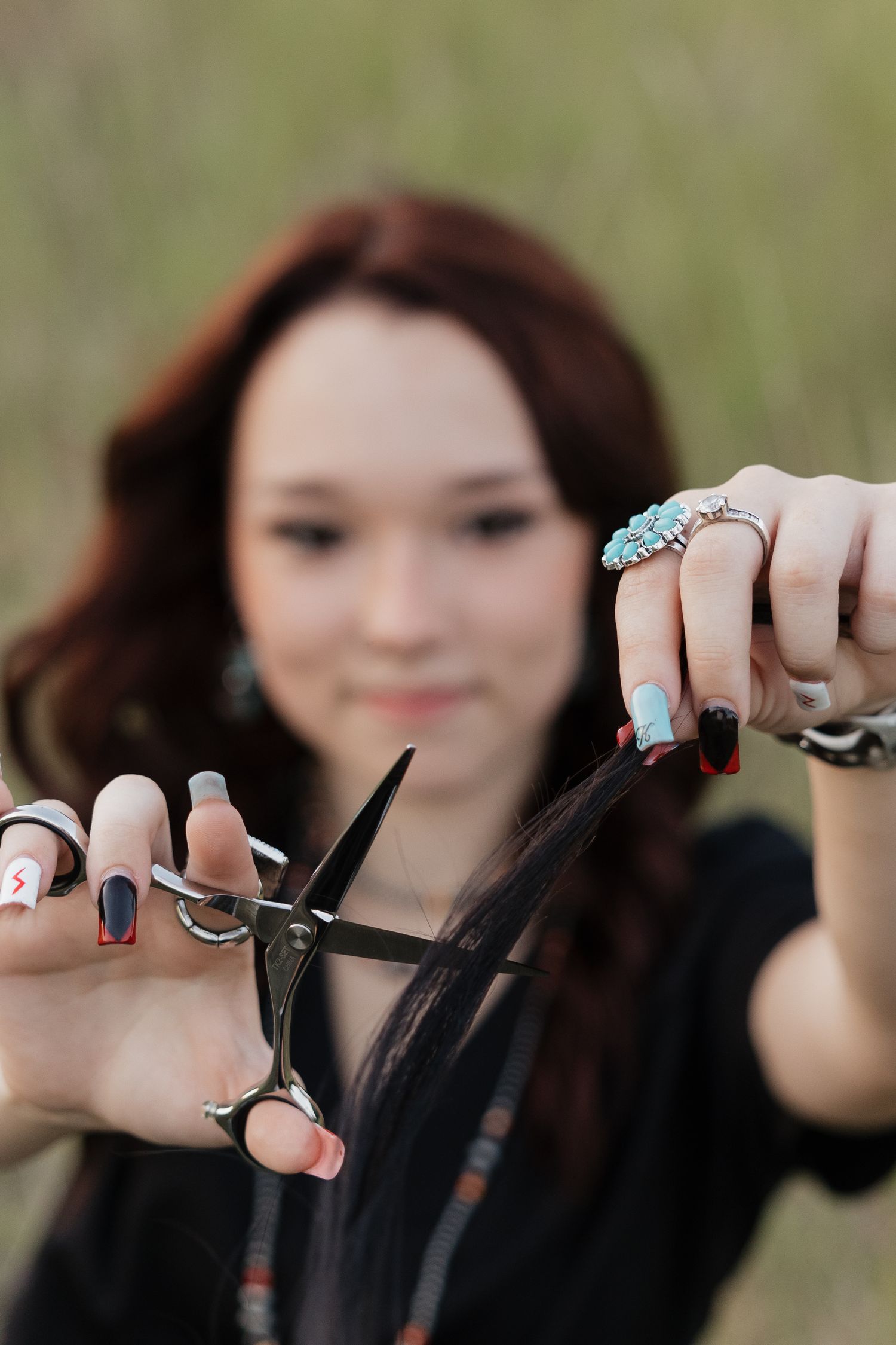 Young woman holding scissors and a lock of dark hair in a grassy outdoor setting with colorful nail art.