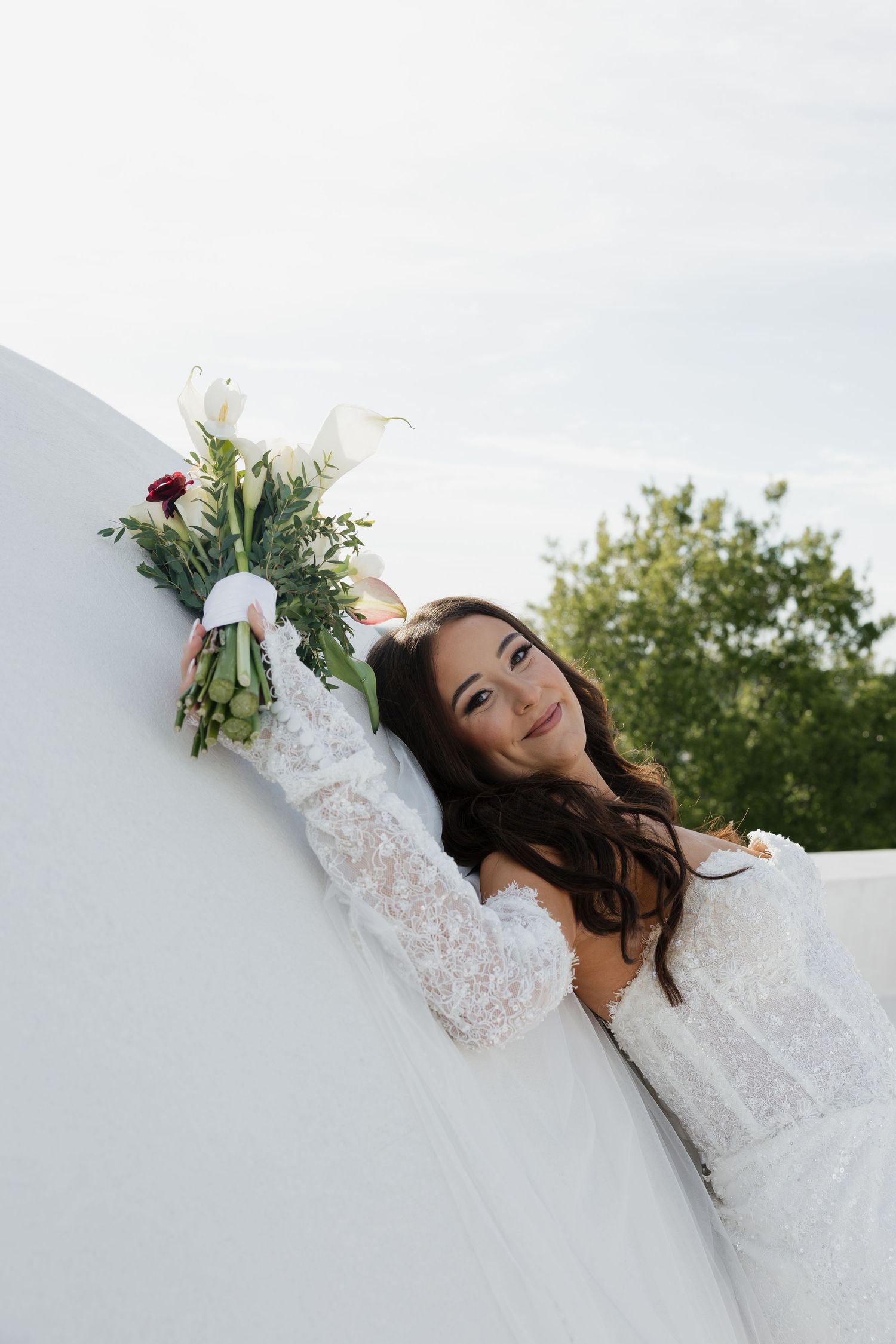 Bride in lace wedding dress leaning against white wall holding floral bouquet with greenery, smiling outdoors.