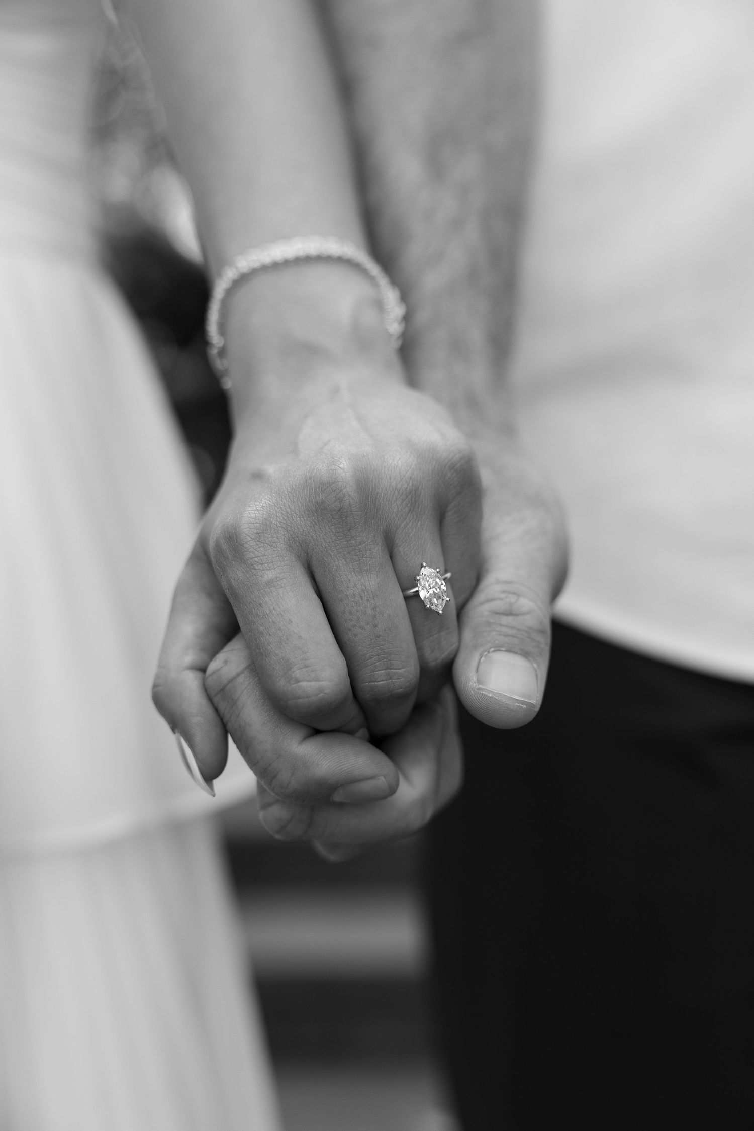 Black and white photo of couple holding hands, showcasing elegant diamond engagement ring and pearl bracelet.