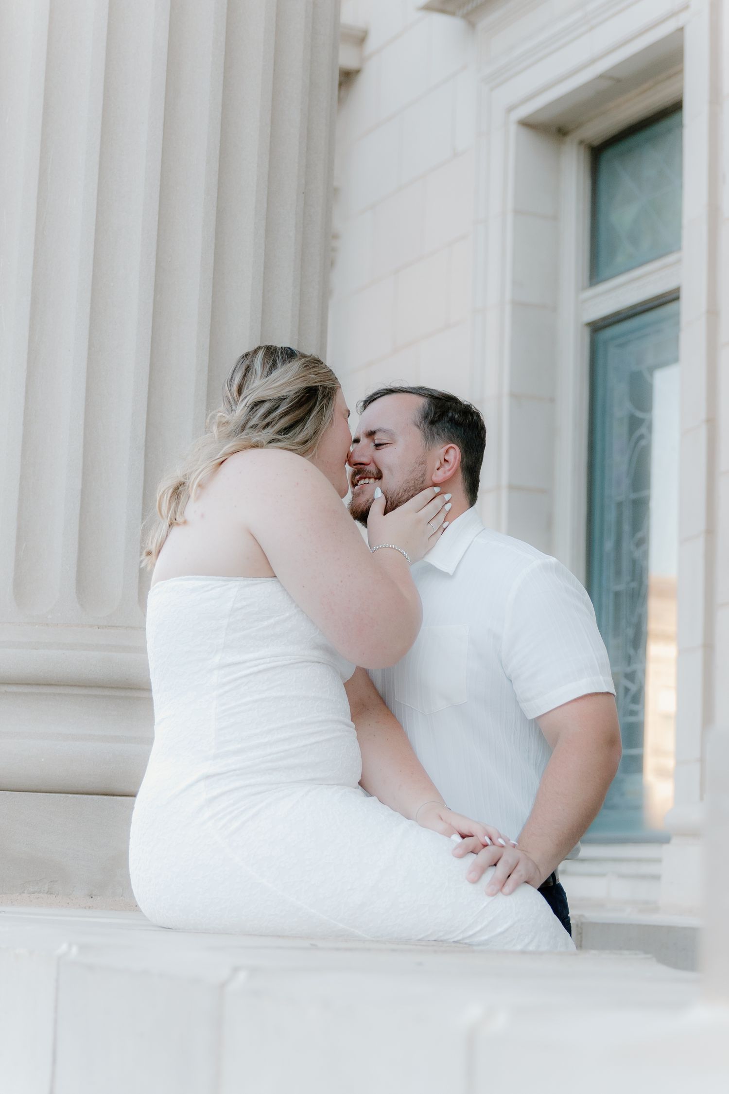 Couple sharing a romantic moment seated on stone columns outside a classical white building.