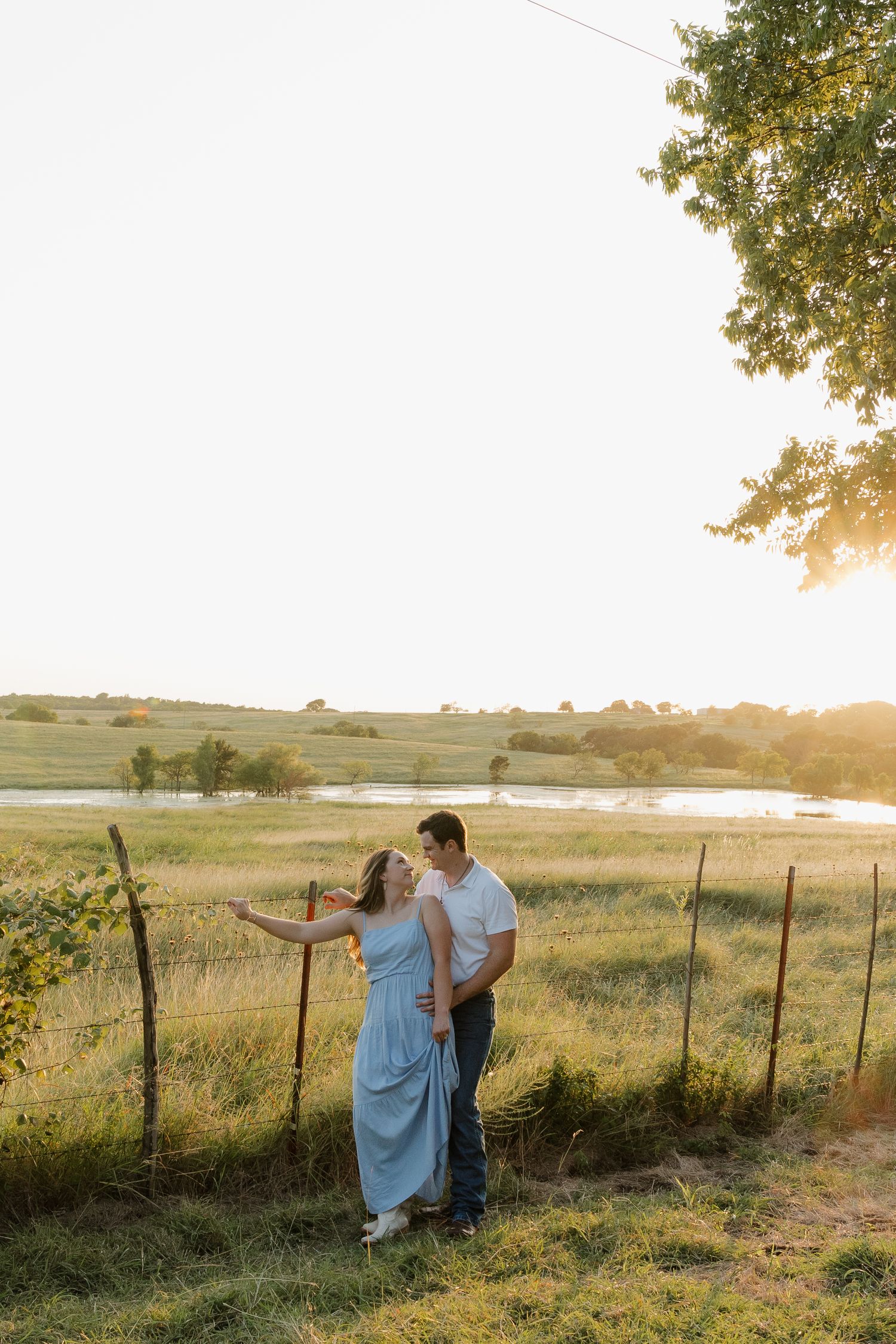 Couple embracing near a rustic fence at golden hour with a scenic pond and rolling fields.