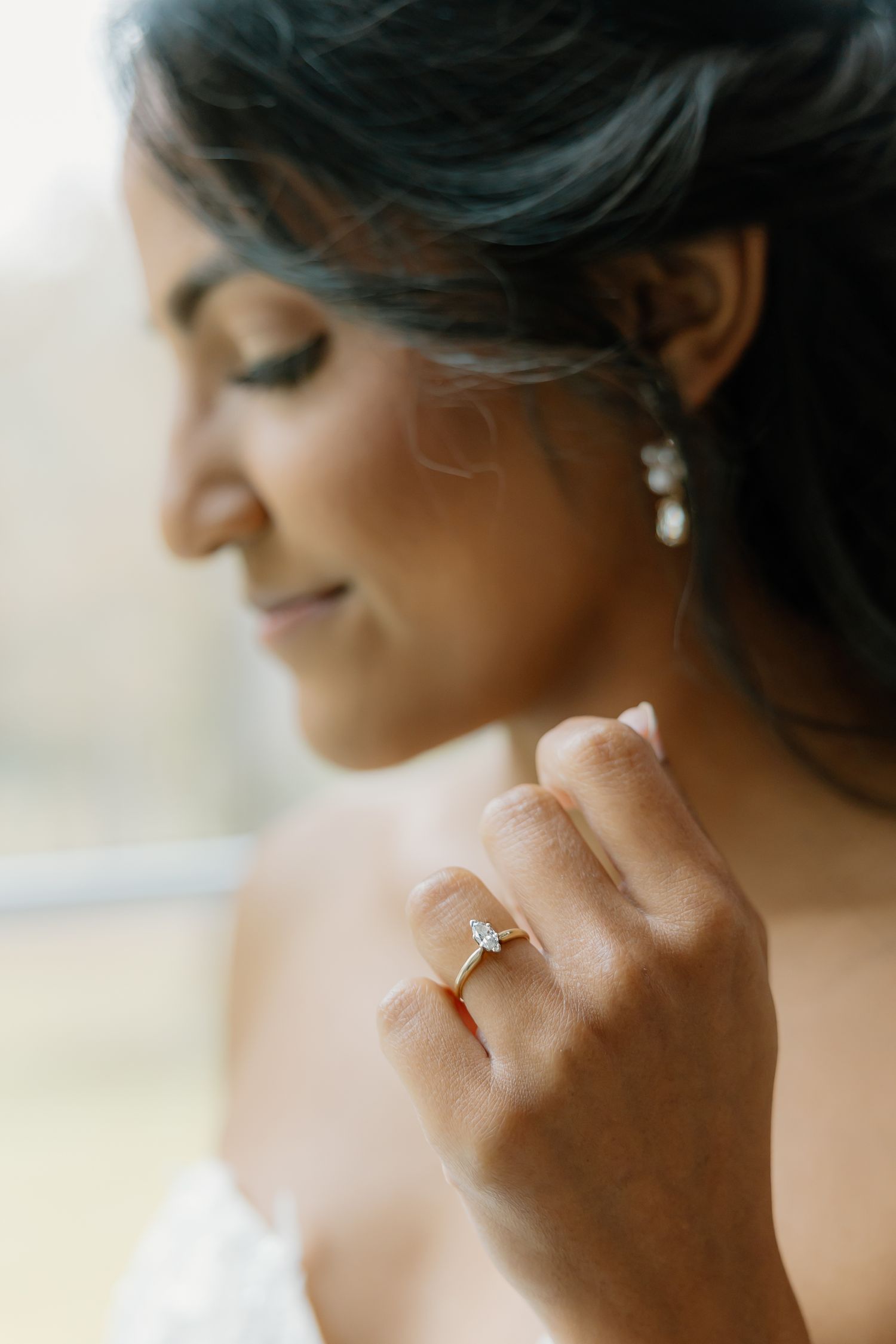 Bride gazes down showcasing elegant diamond engagement ring on gold band with pearl drop earrings.