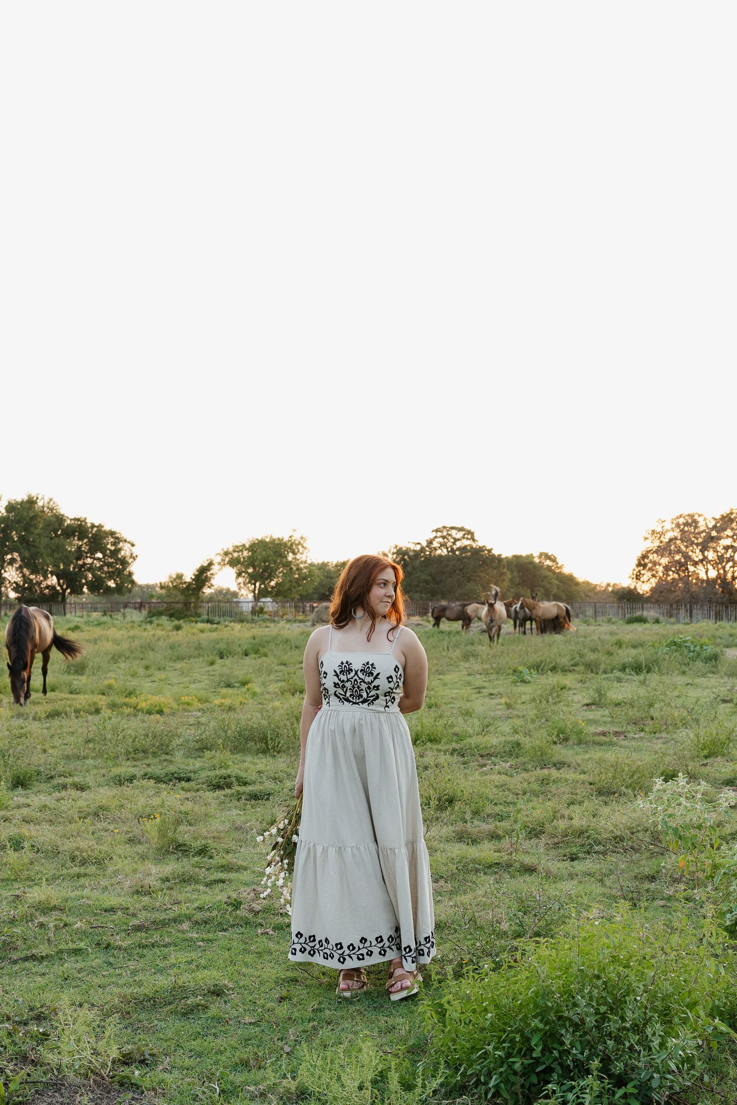 Woman in embroidered white maxi dress holding wildflowers in a green pasture.