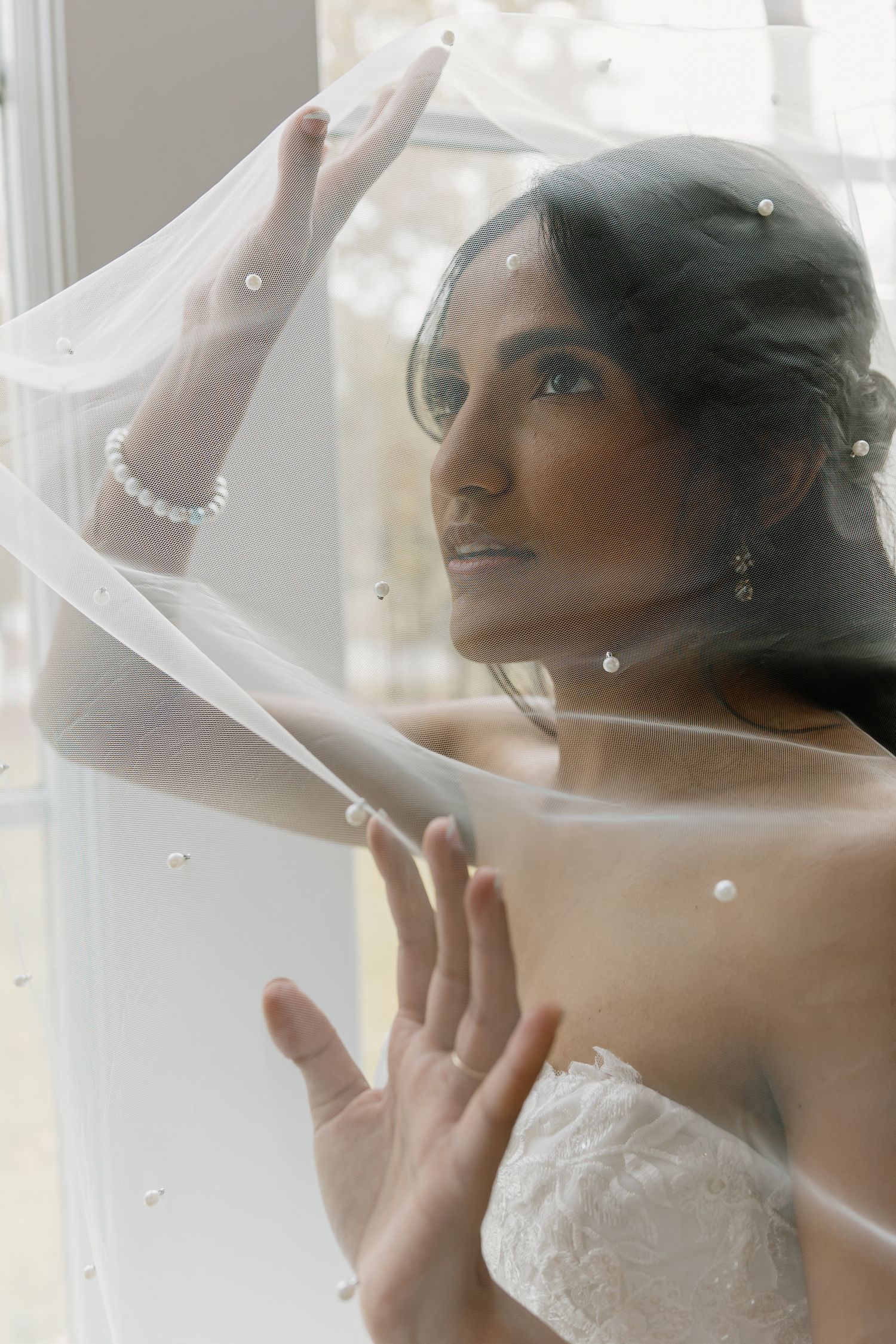 Beautiful bride holding veil near window light wearing pearl bracelet and strapless lace gown.