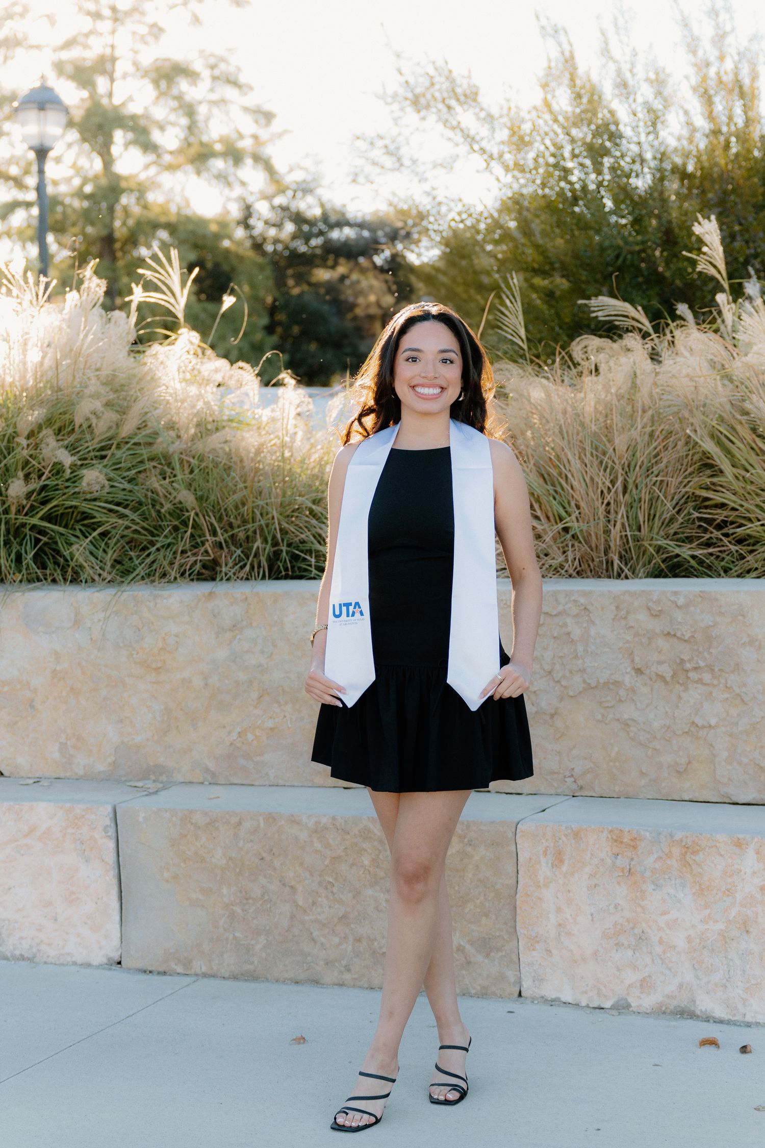 Smiling UTA graduate in black dress with white graduation stole standing outdoors among ornamental grasses.