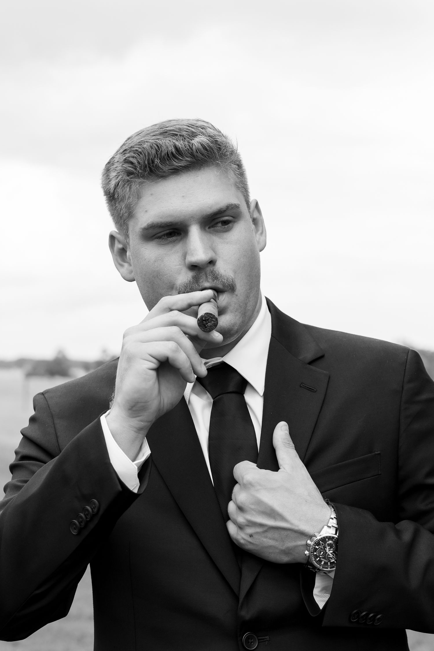 Dapper groom in black suit and tie smoking a cigar in dramatic black and white portrait.
