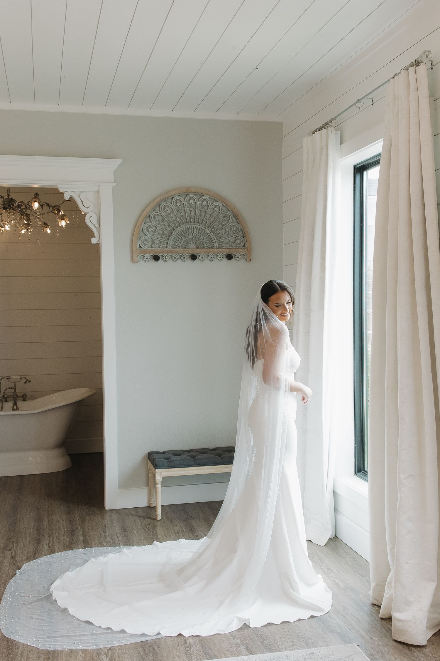 Bride in white gown and long veil stands by window in elegant bridal suite with clawfoot tub.