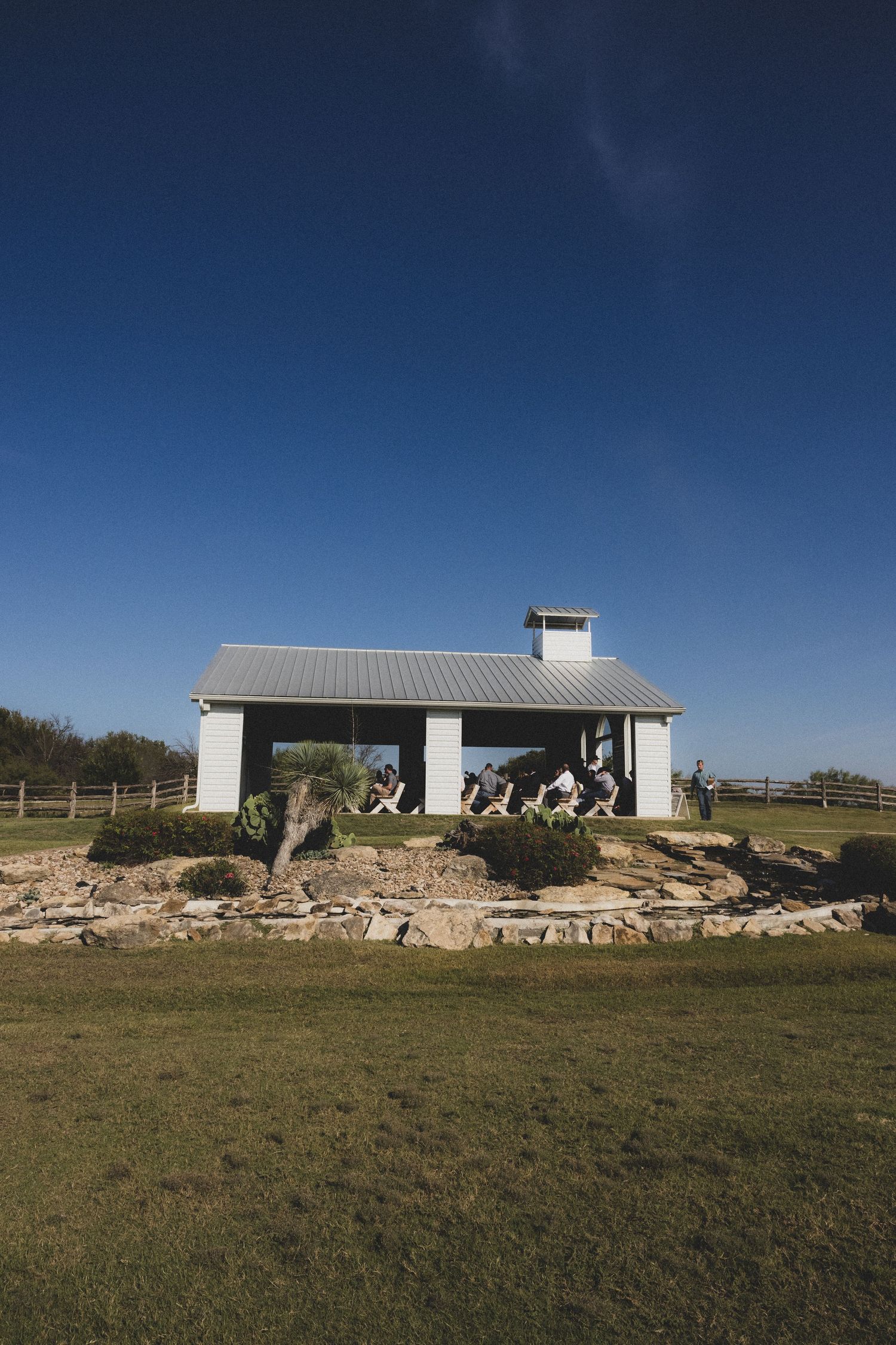 White farmhouse wedding venue with metal roof surrounded by green grass under a deep blue sky.