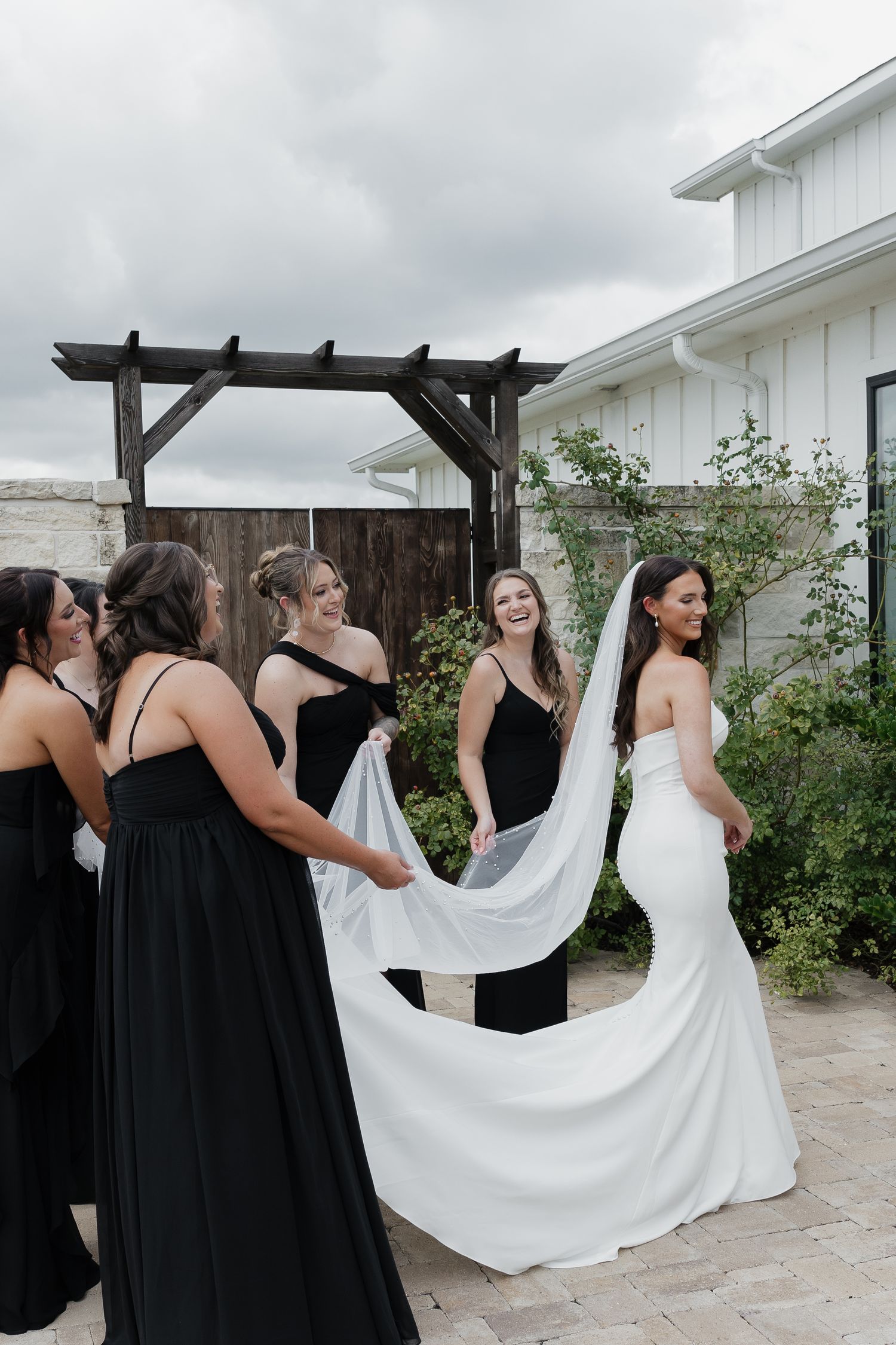 Bridesmaids in black gowns help carry brides long white veil and train outside white barn venue.