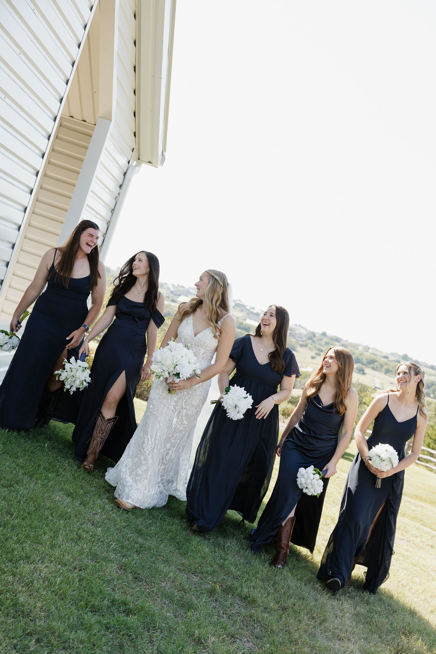 Bride in lace gown walks with bridesmaids in navy dresses holding white bouquets outdoors.