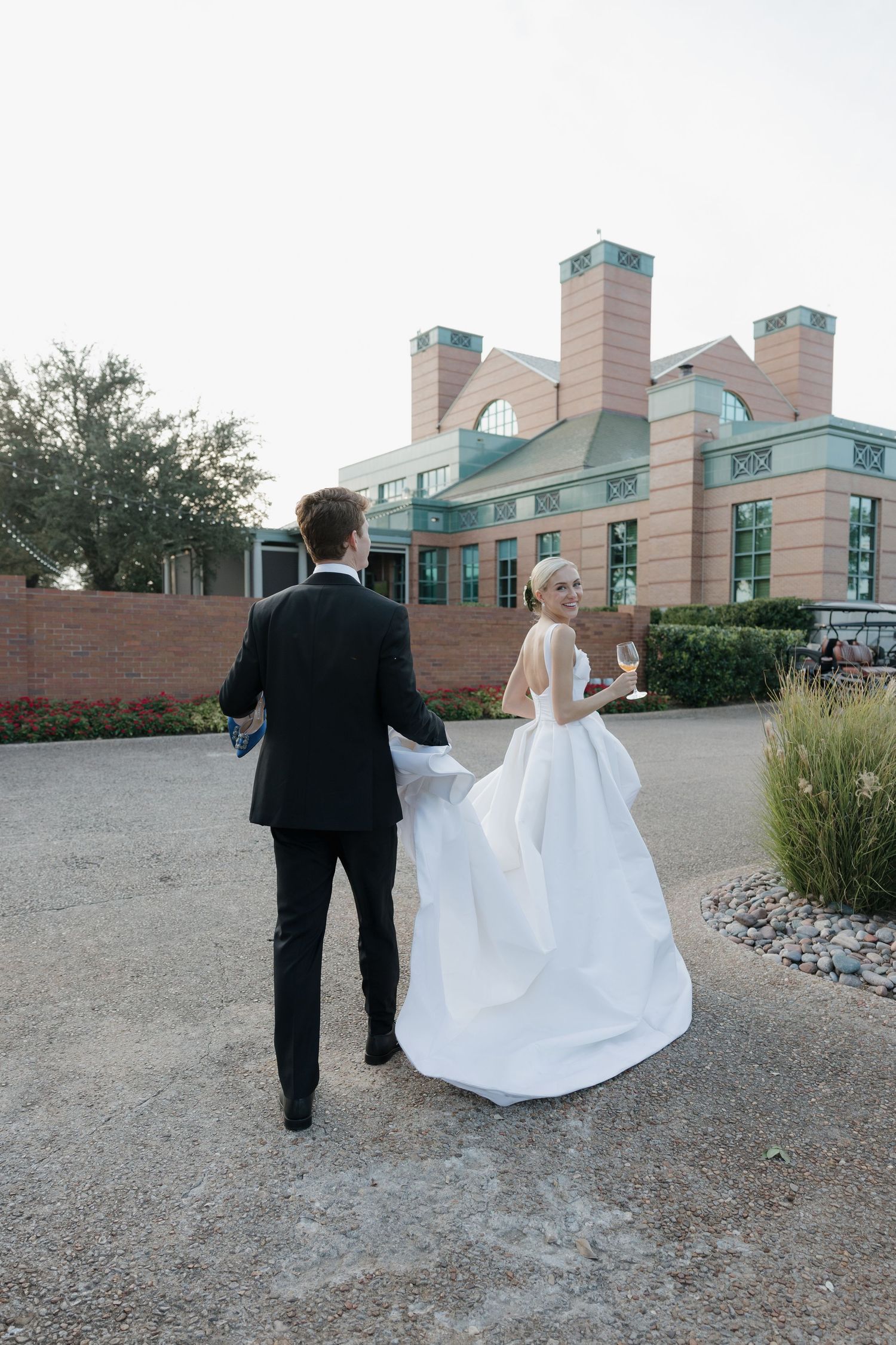 Wedding couple strolling toward brick venue, bride in white ball gown holding champagne glass, groom in black tuxedo.