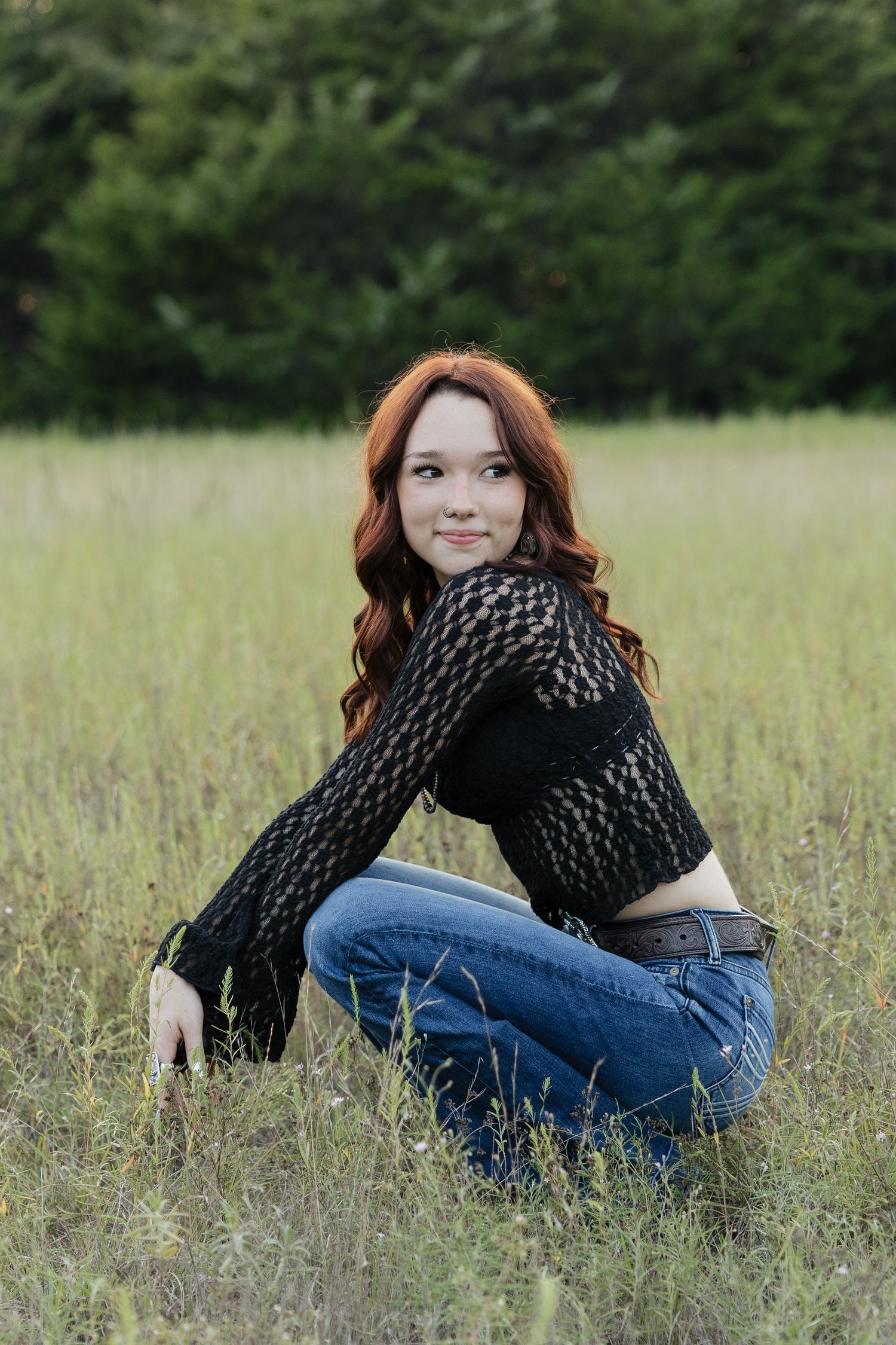 Young woman with red wavy hair in a black lace top and jeans crouching in a grassy field smiling.