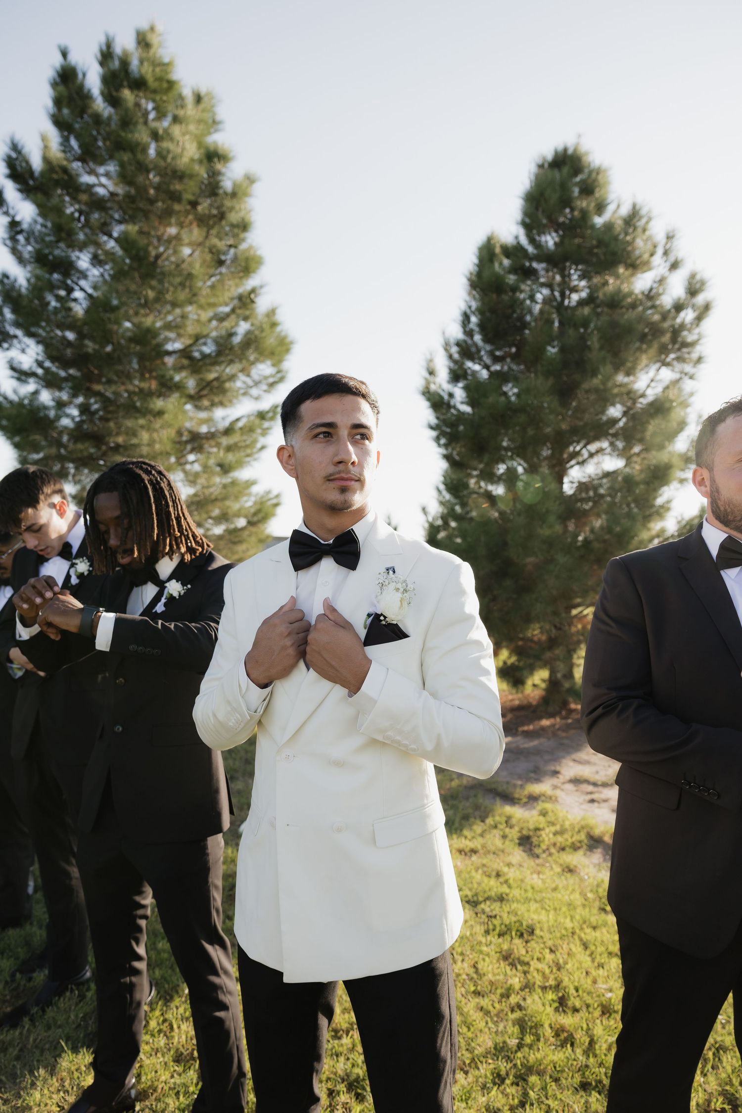Handsome groom in white tuxedo jacket adjusts bow tie surrounded by groomsmen outdoors.