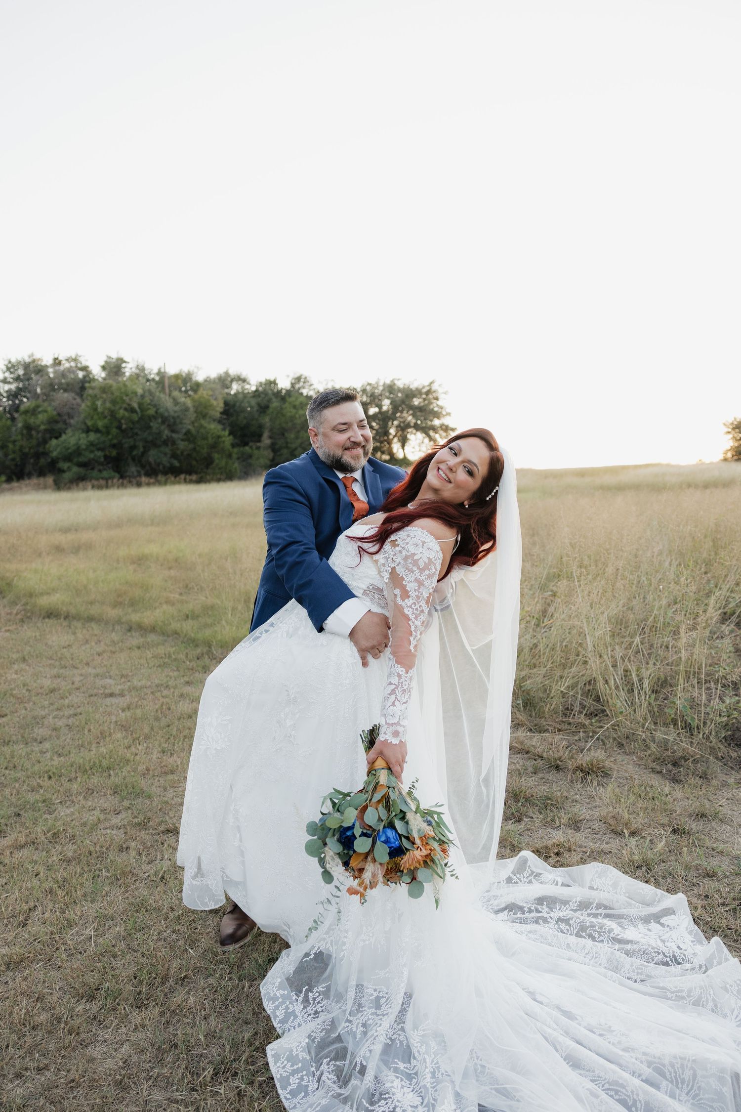 Groom lifts smiling bride in lace gown and veil in golden hour field holding floral bouquet.