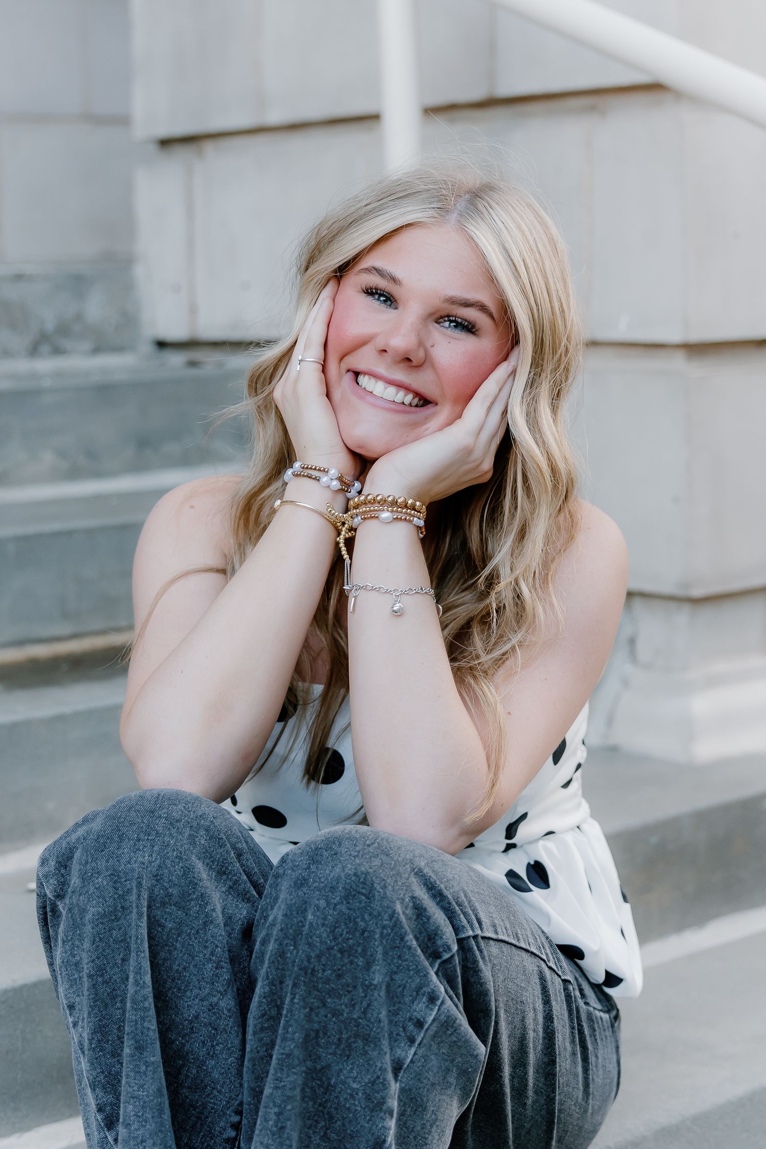 Smiling blonde teen girl in polka dot top sitting on stone steps with hands on cheeks wearing gold bracelets.