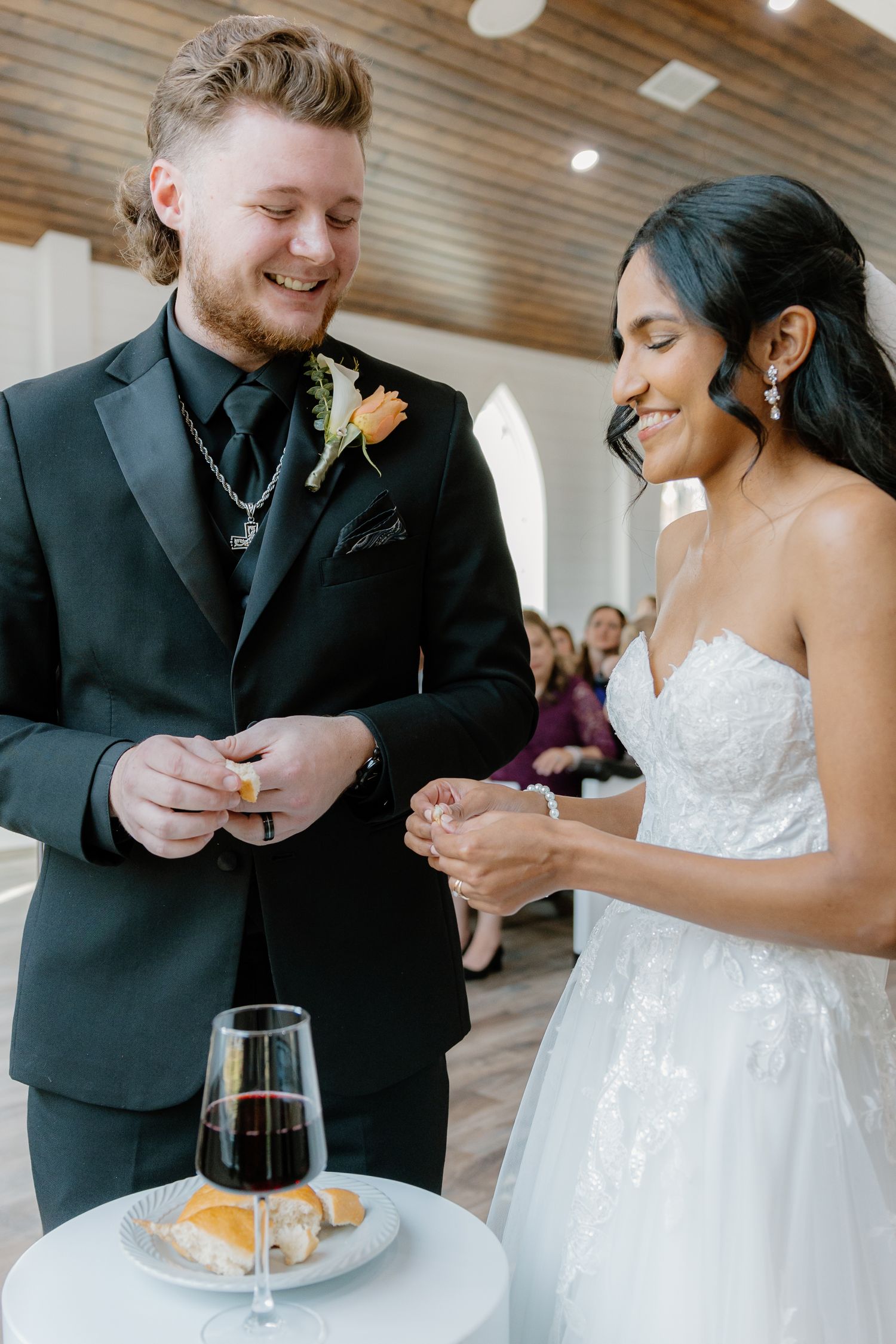 Smiling bride and groom sharing communion bread and wine during indoor wedding ceremony.