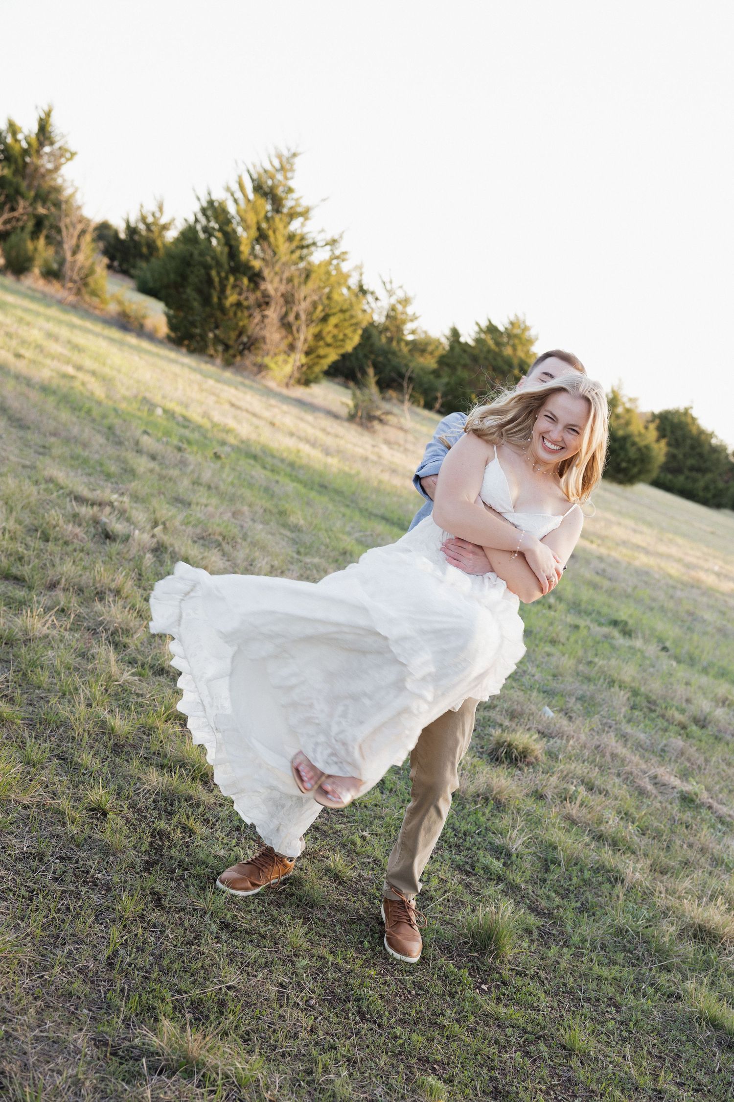 Groom lifting bride in white dress outdoors in a sunny green field, both smiling joyfully during wedding photos.