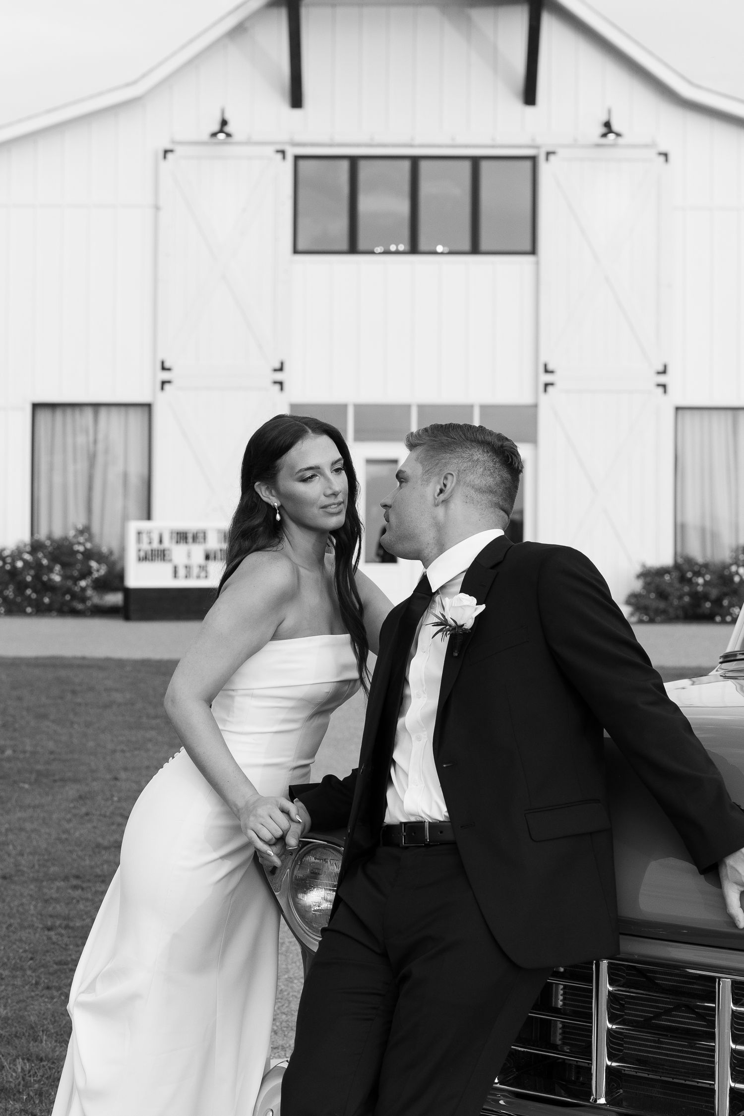 Black and white photo of bride and groom leaning against vintage car in front of white barn.