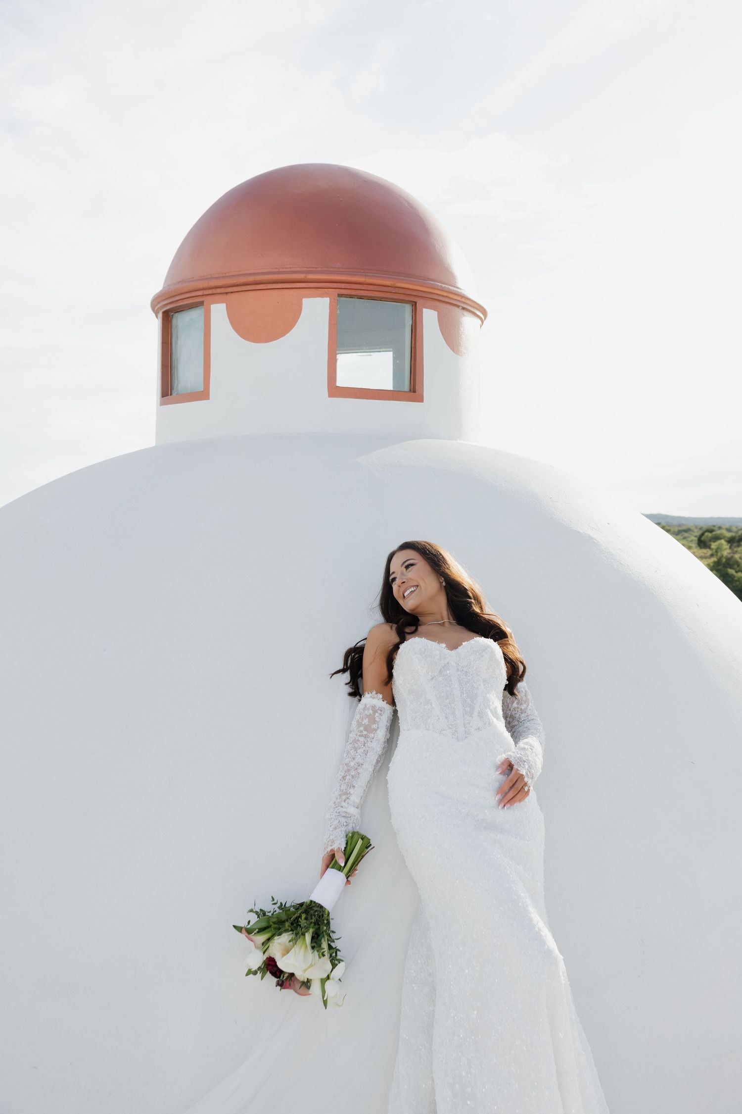 Bride in white lace off-shoulder gown holding bouquet, posing against white domed building with terracotta roof.
